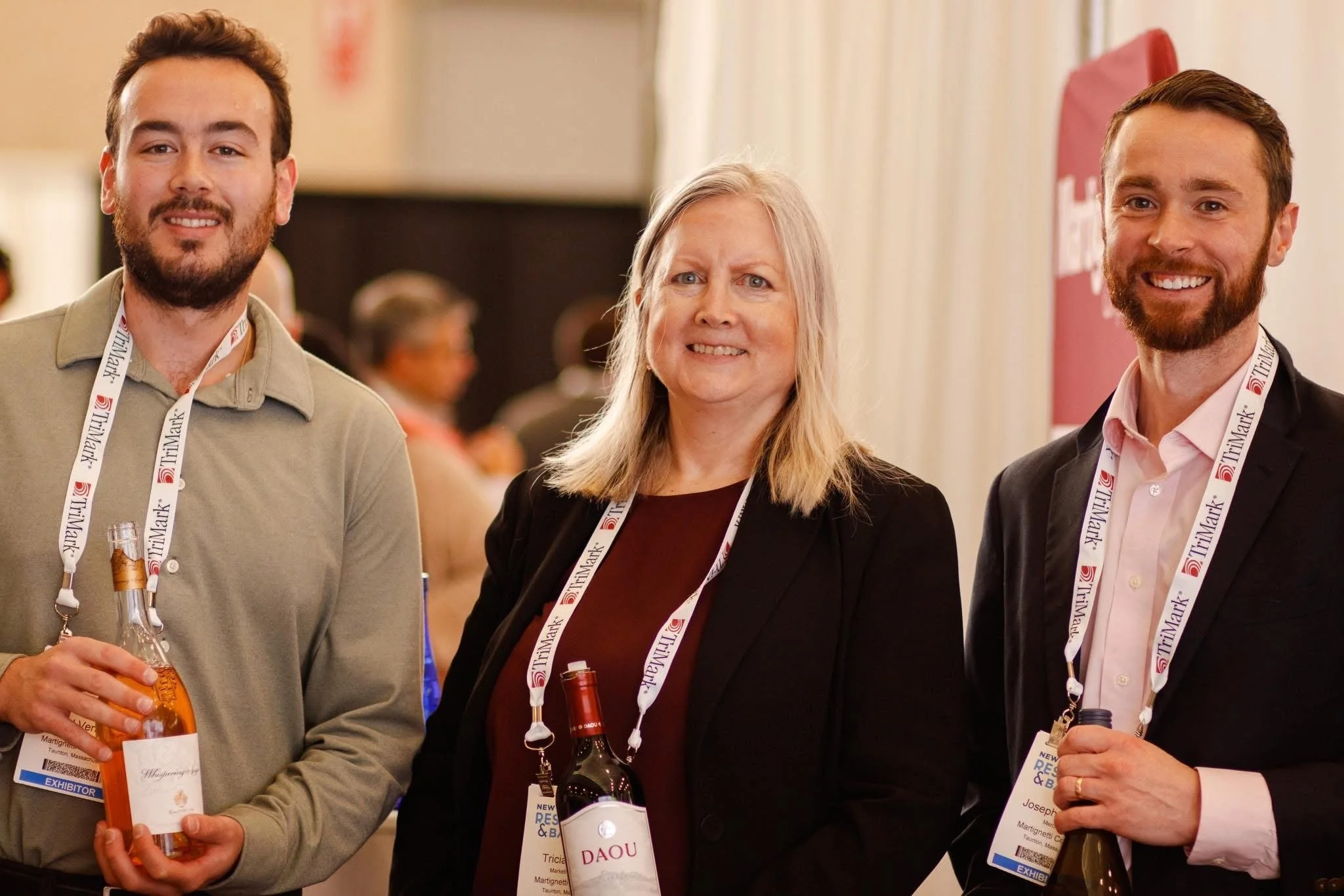 Three people at a conference or event, smiling and holding drinks, wearing badges around their necks.