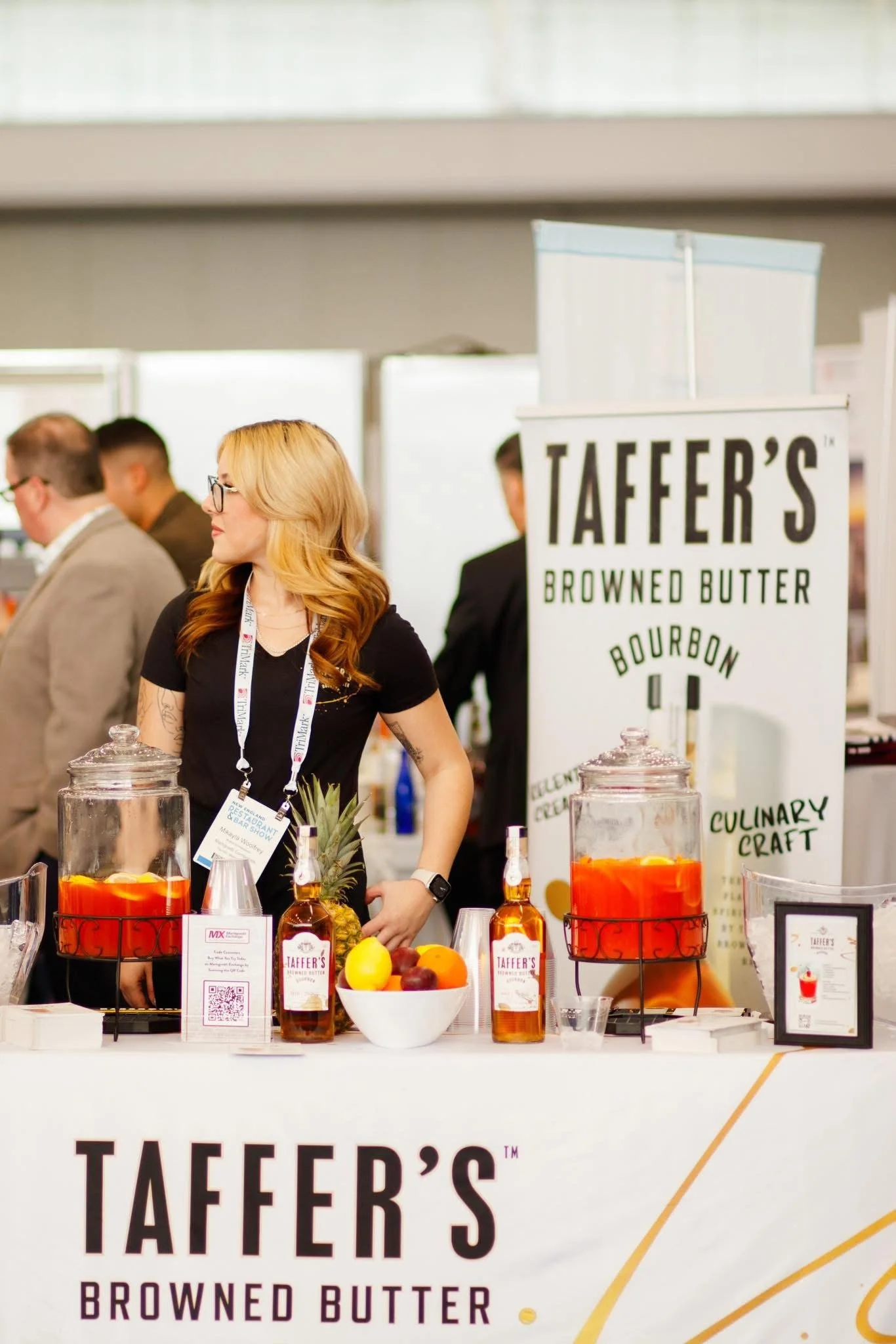 A woman with blonde hair and glasses standing behind a booth at a food or beverage event, with various bottles and a pineapple on display, and a sign that reads 'Taffer's Browned Butter Bourbon'.