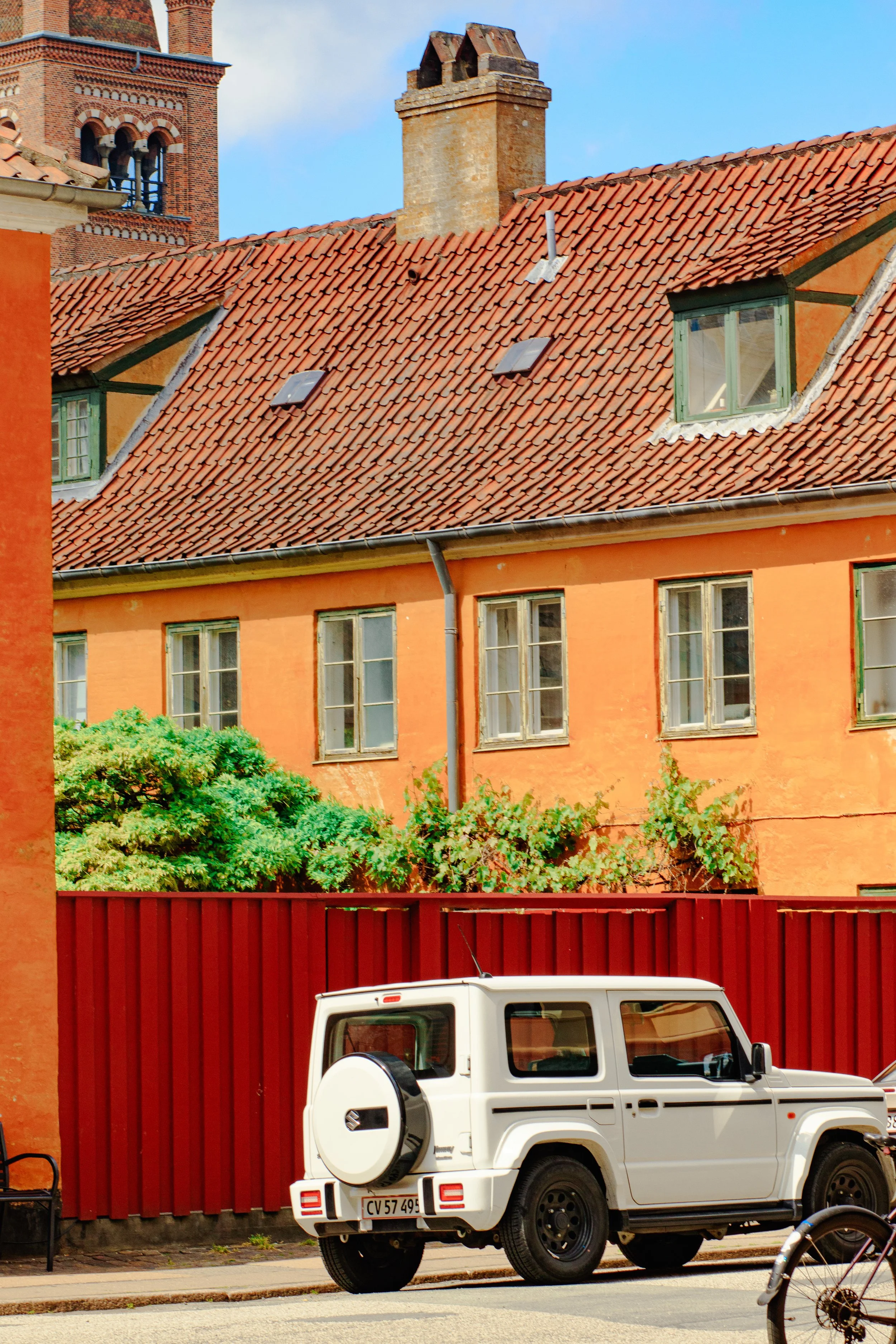 A white SUV parked in front of a red wooden fence and a peach-colored building with multiple windows, green window frames, and a tiled roof, with a lush green bush below the windows.