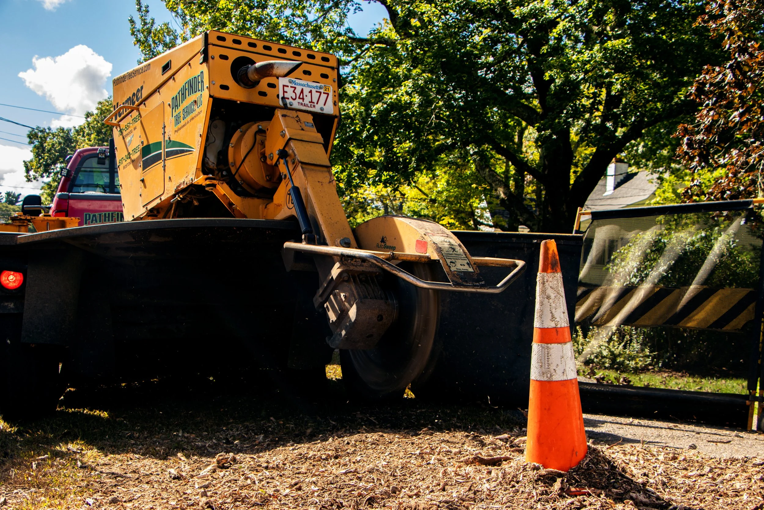 A construction vehicle parked outdoors with an orange and white traffic cone in the foreground. The vehicle is yellow with equipment on the back, and there's a tree and a house in the background.