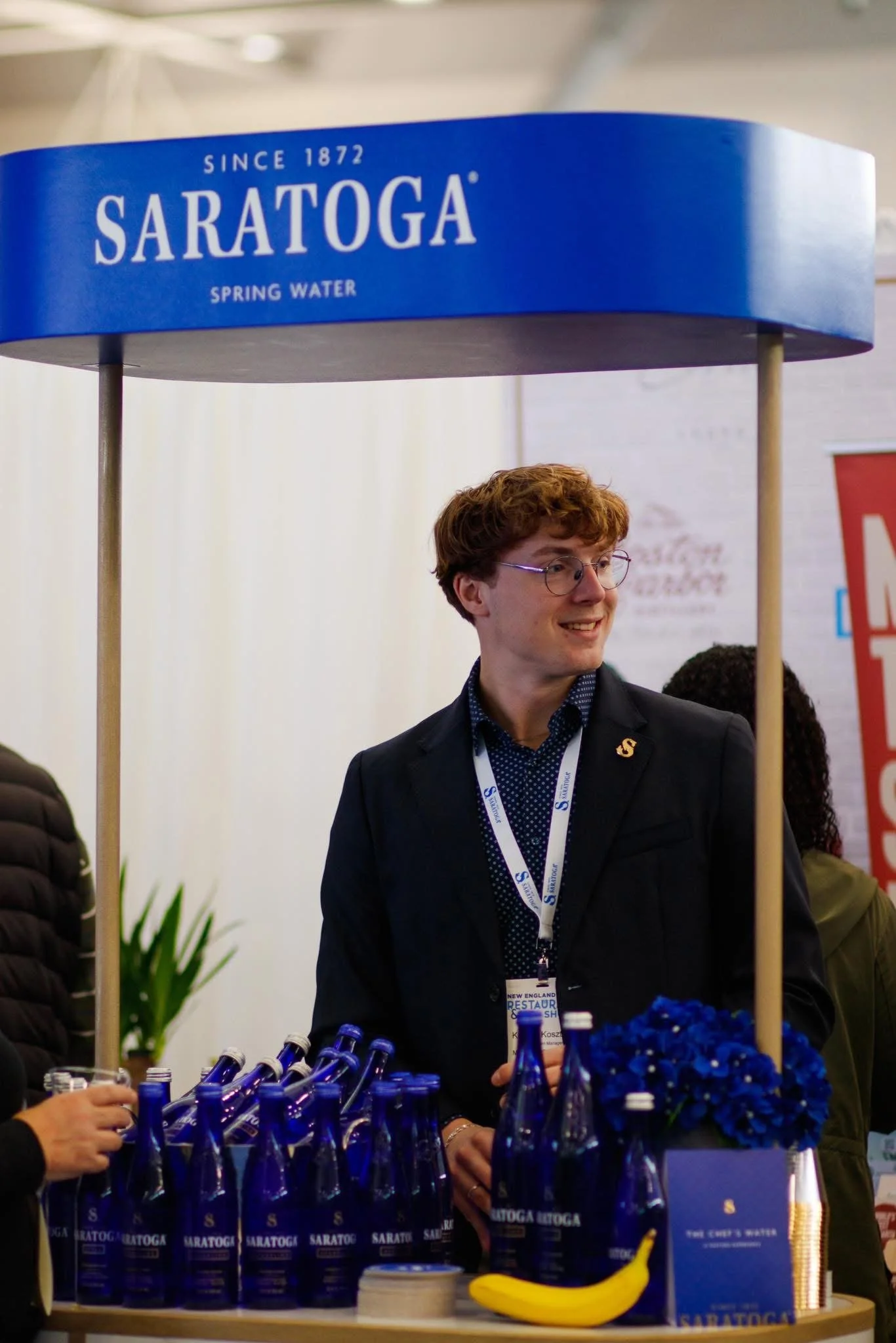 A man with glasses and a dark suit standing behind a booth at an event with a sign that says "Saratoga Spring Water Since 1872." The booth has blue water bottles labeled "Saratoga." There are blue flowers and a banana on the counter, and people are a