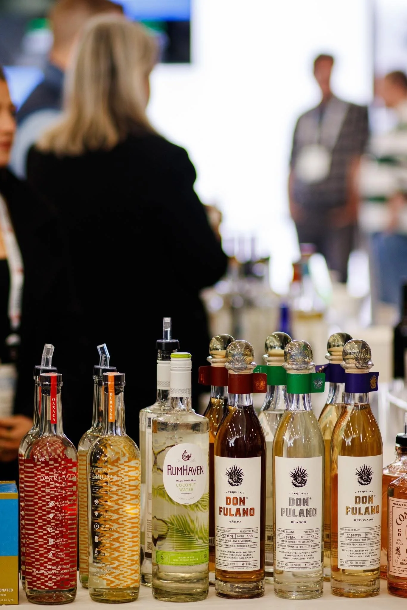 Various bottles of alcohol on display in a store, with blurred people in the background.