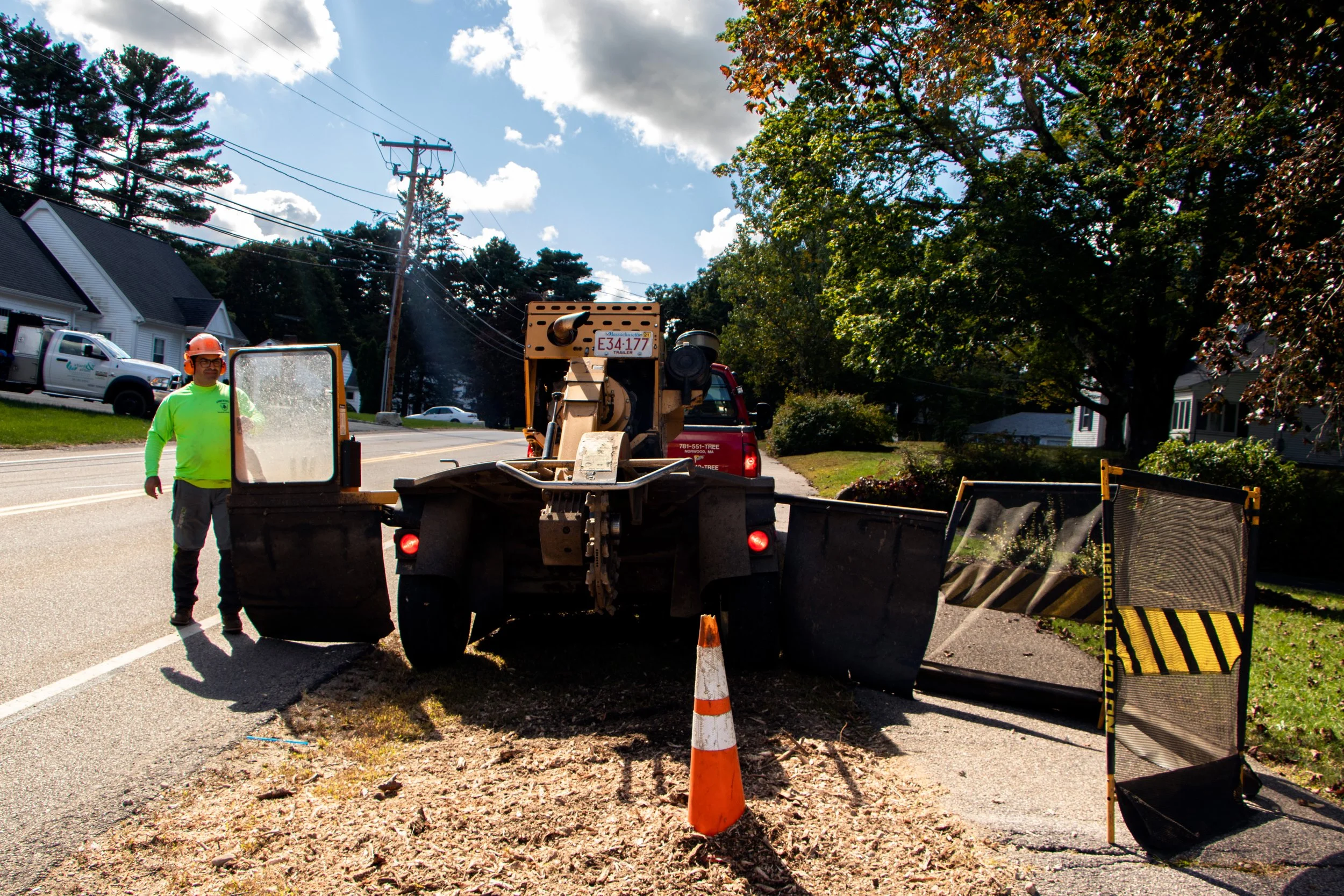 A man in a neon yellow shirt and orange hard hat working on a street construction site. There is a large piece of construction equipment and traffic cones, with trees, houses, and utility poles in the background.