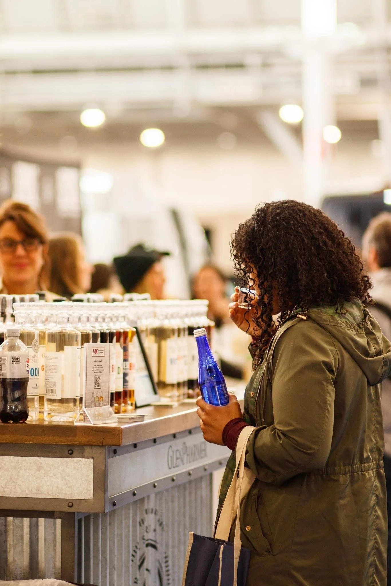 A woman with curly hair wearing an olive green jacket shopping at a store, holding a blue glass bottle, with a row of bottles and sign on the counter in front of her, and several women and a man in the background.