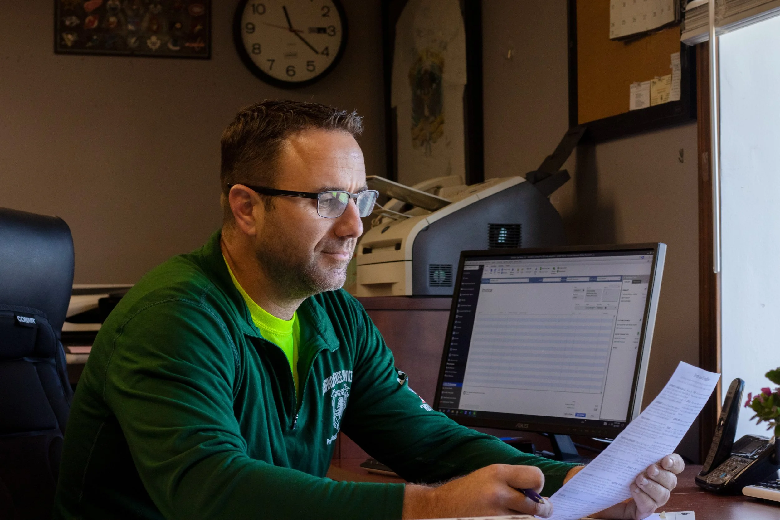 A man wearing glasses and a green jacket sits at a desk, reading a sheet of paper. In front of him is a computer monitor displaying a spreadsheet. The background features a clock, framed artwork, a bulletin board, and office supplies.