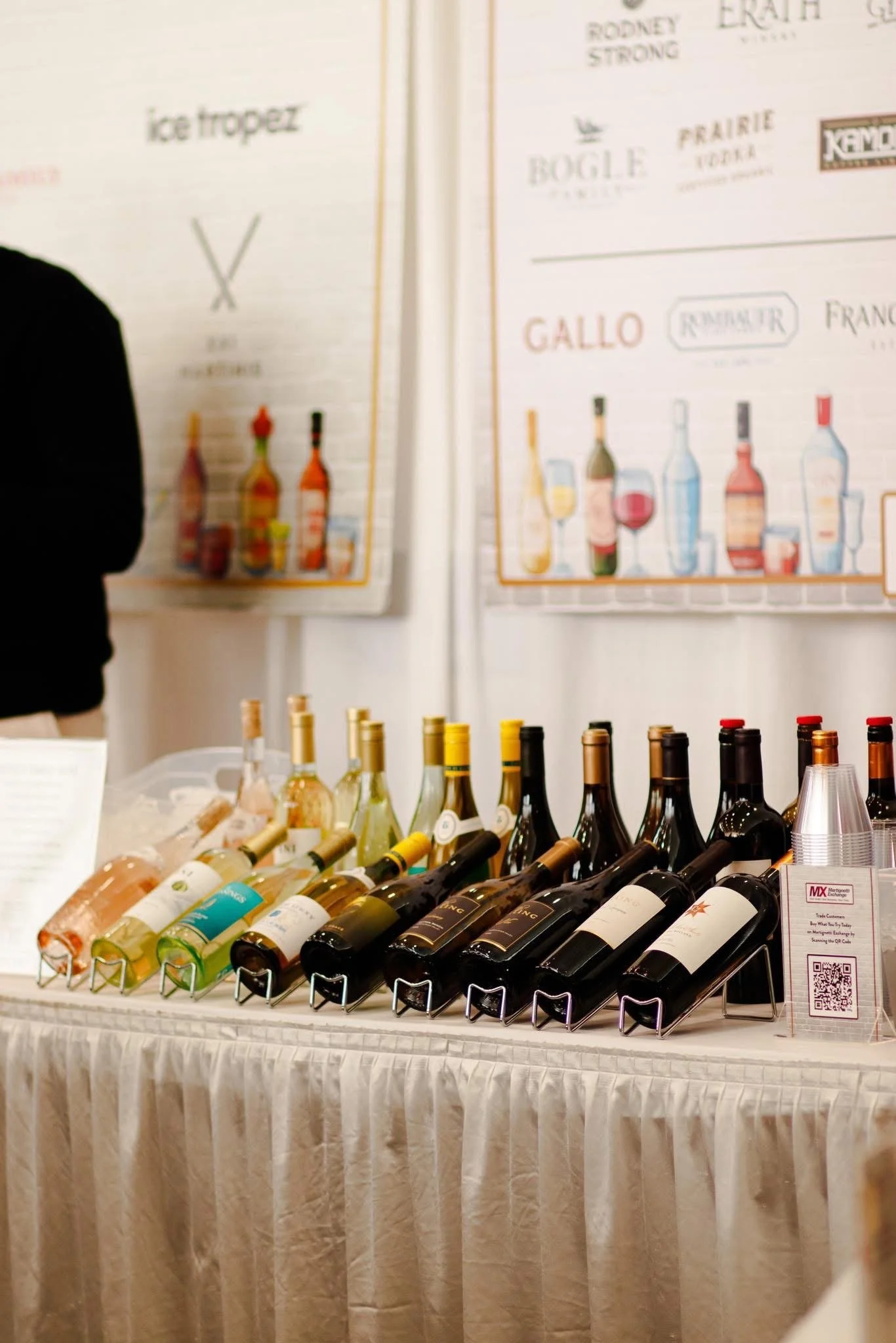 A table with various bottles of wine arranged in rows, a bottled water, and a QR code sign, with wine-related posters on the wall in the background.