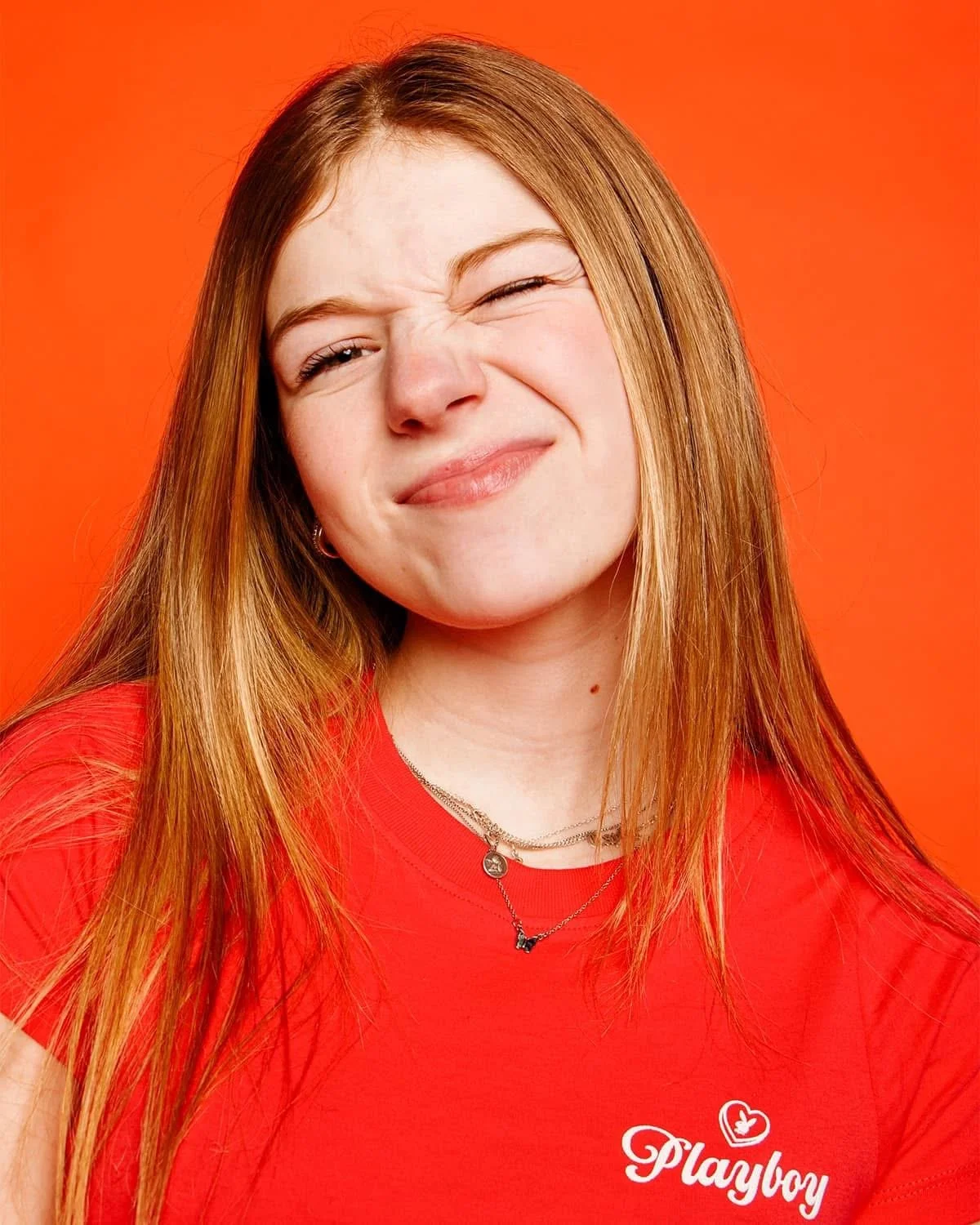 A woman with long red hair wearing a red Playbuzz t-shirt against an orange background, making a playful facial expression with one eye closed and mouth slightly twisted.
