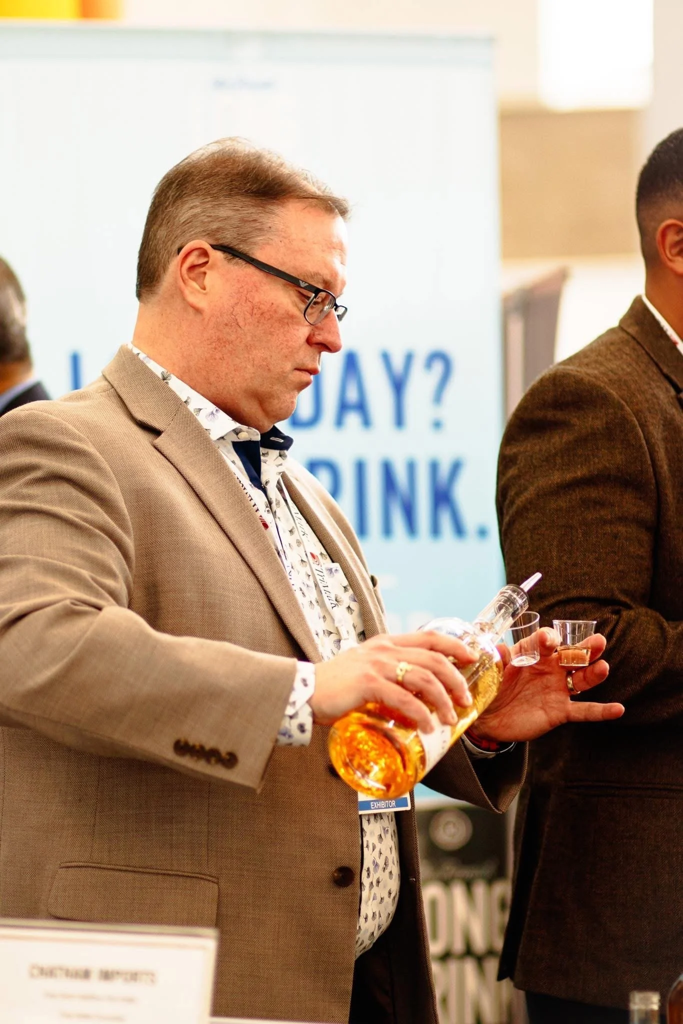 A man in a beige blazer and glasses pouring a drink from a bottle into a small glass at a public event.