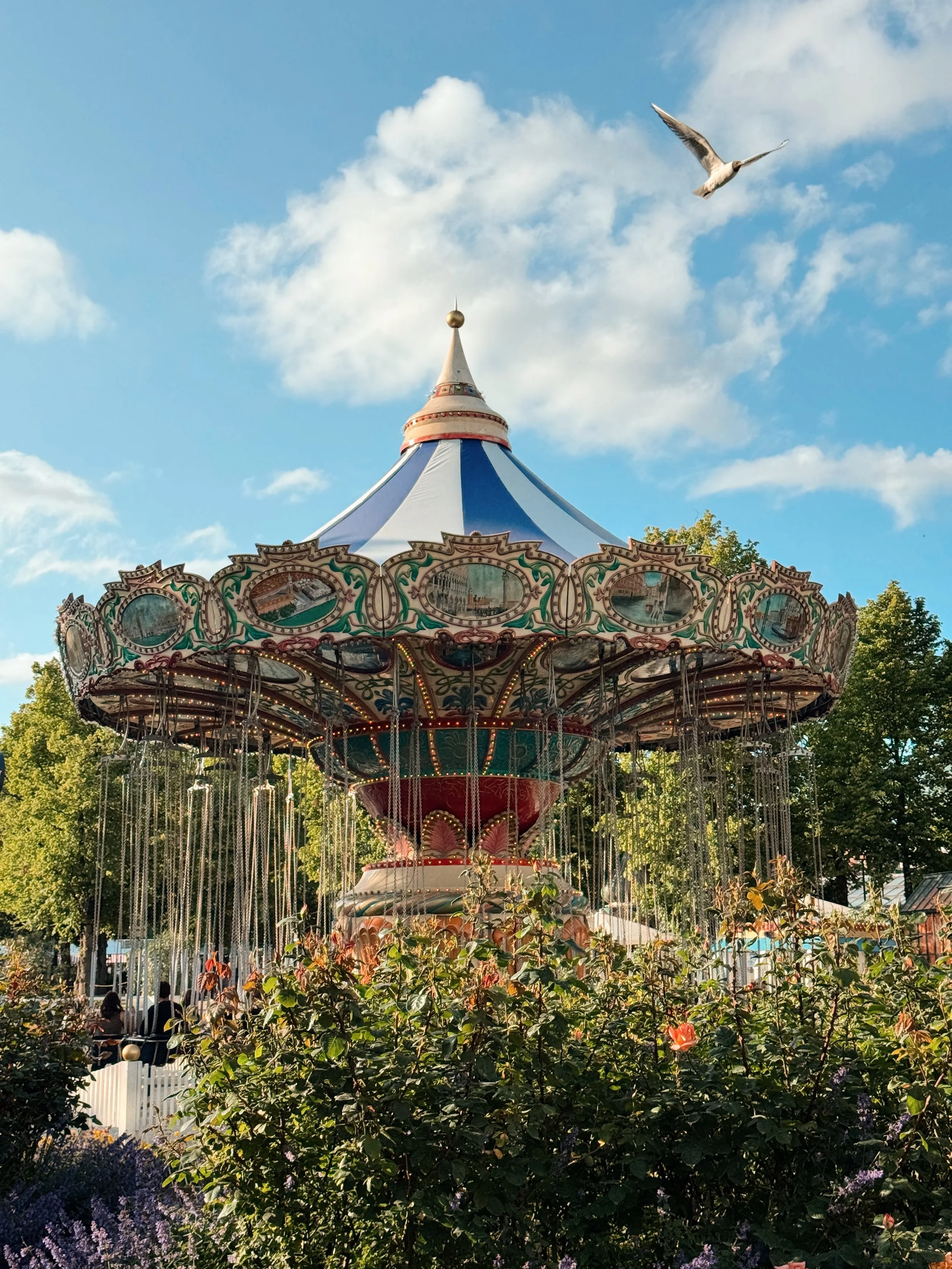 A vintage-style carousel with ornate decorations, featuring a blue and white tent-like top and a golden spire, surrounded by green trees and purple flowers, with people riding it and a seagull flying in the blue sky with scattered clouds.
