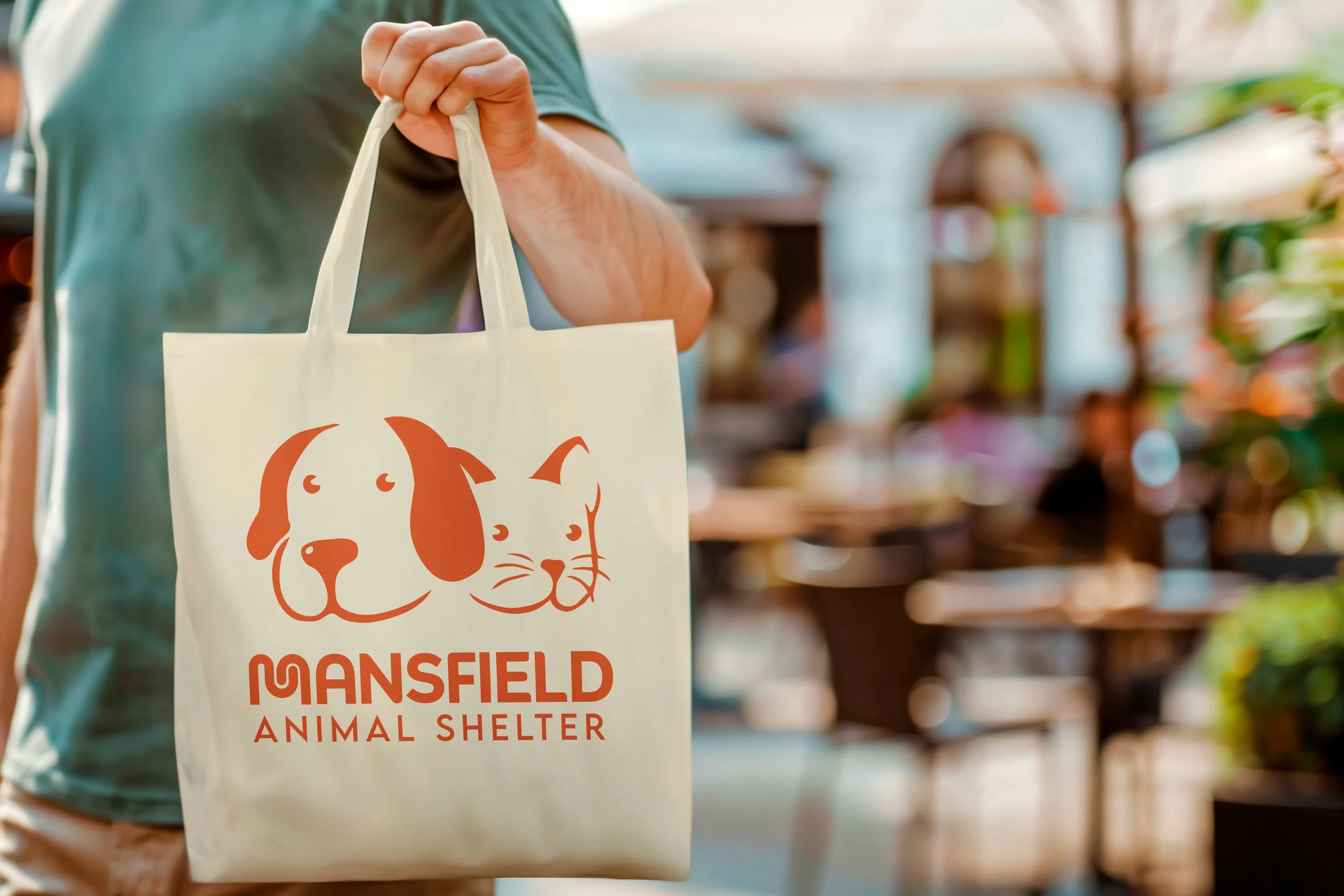 Person holding a donation bag with dog faces and the words Mansfield Animal Shelter printed on it, in a store or similar setting.