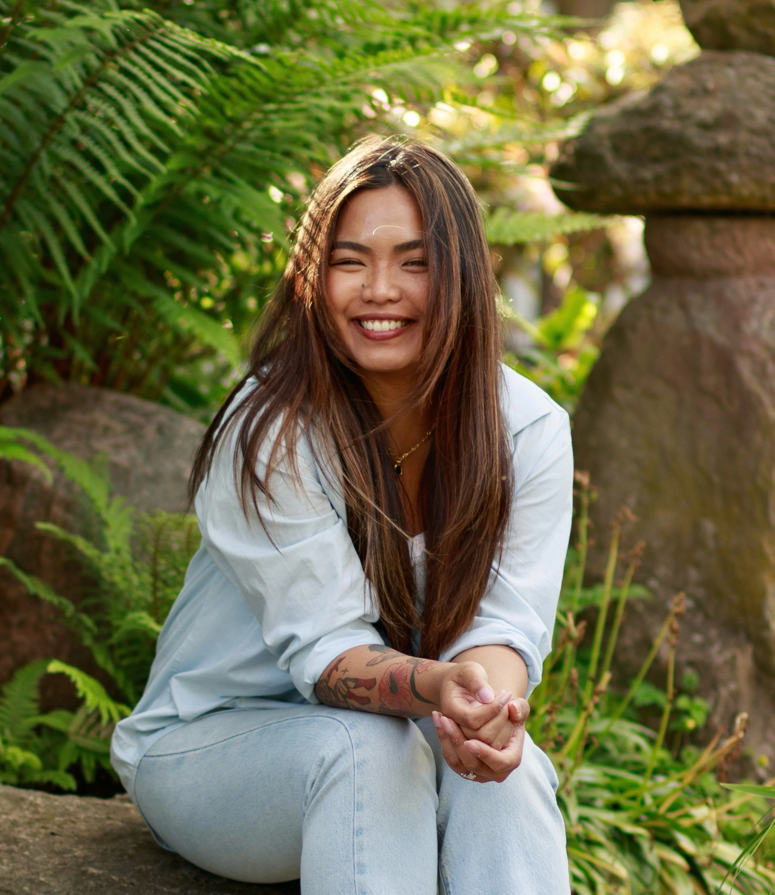 A young woman with long brown hair and tattoos on her arm, smiling and sitting on the ground outdoors surrounded by green ferns and rocks.