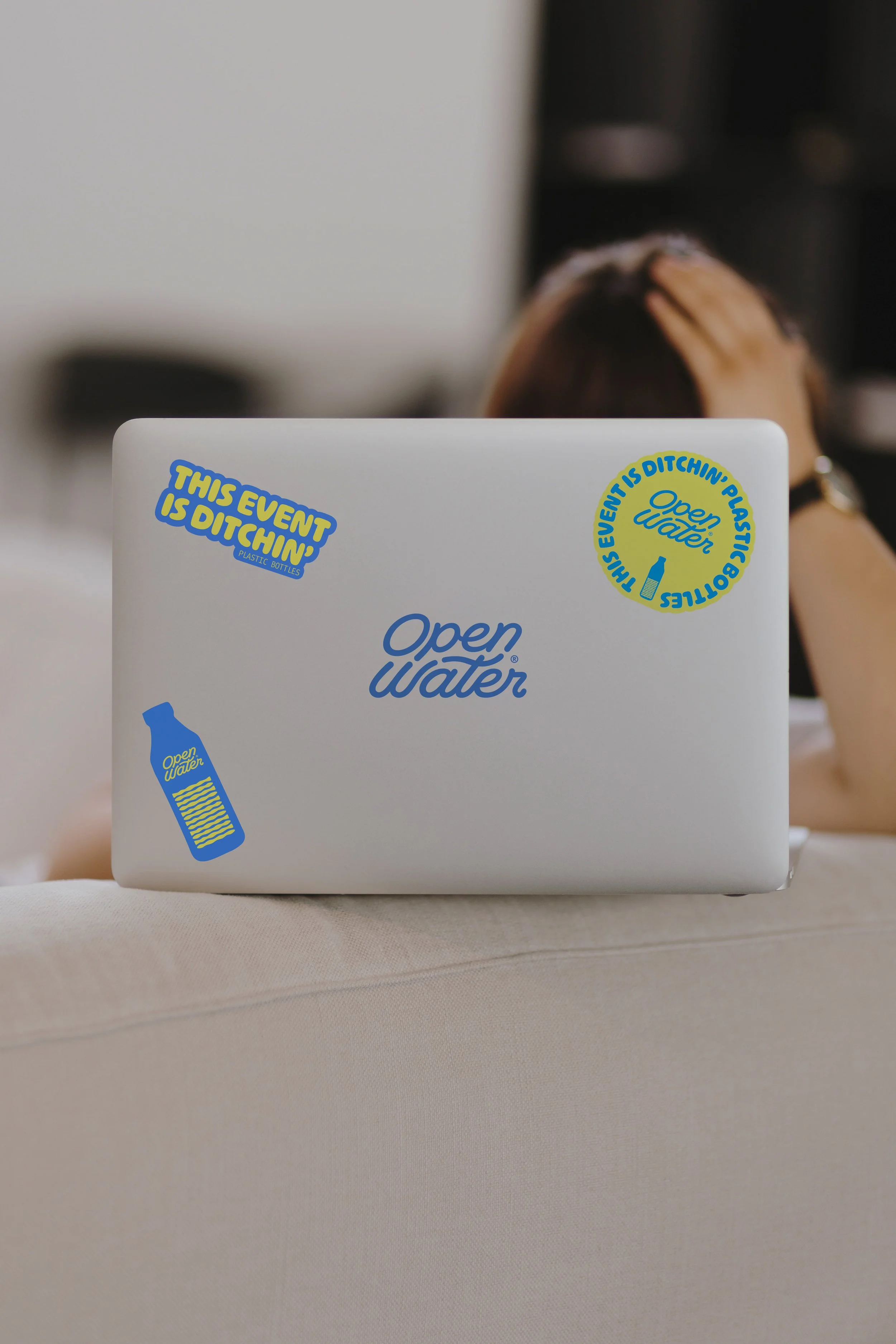 A laptop decorated with water-themed stickers, including one that says "This event is Dutchin'" and another that says "Open Water" with a bottle icon. A person with brown hair is partially visible in the background.