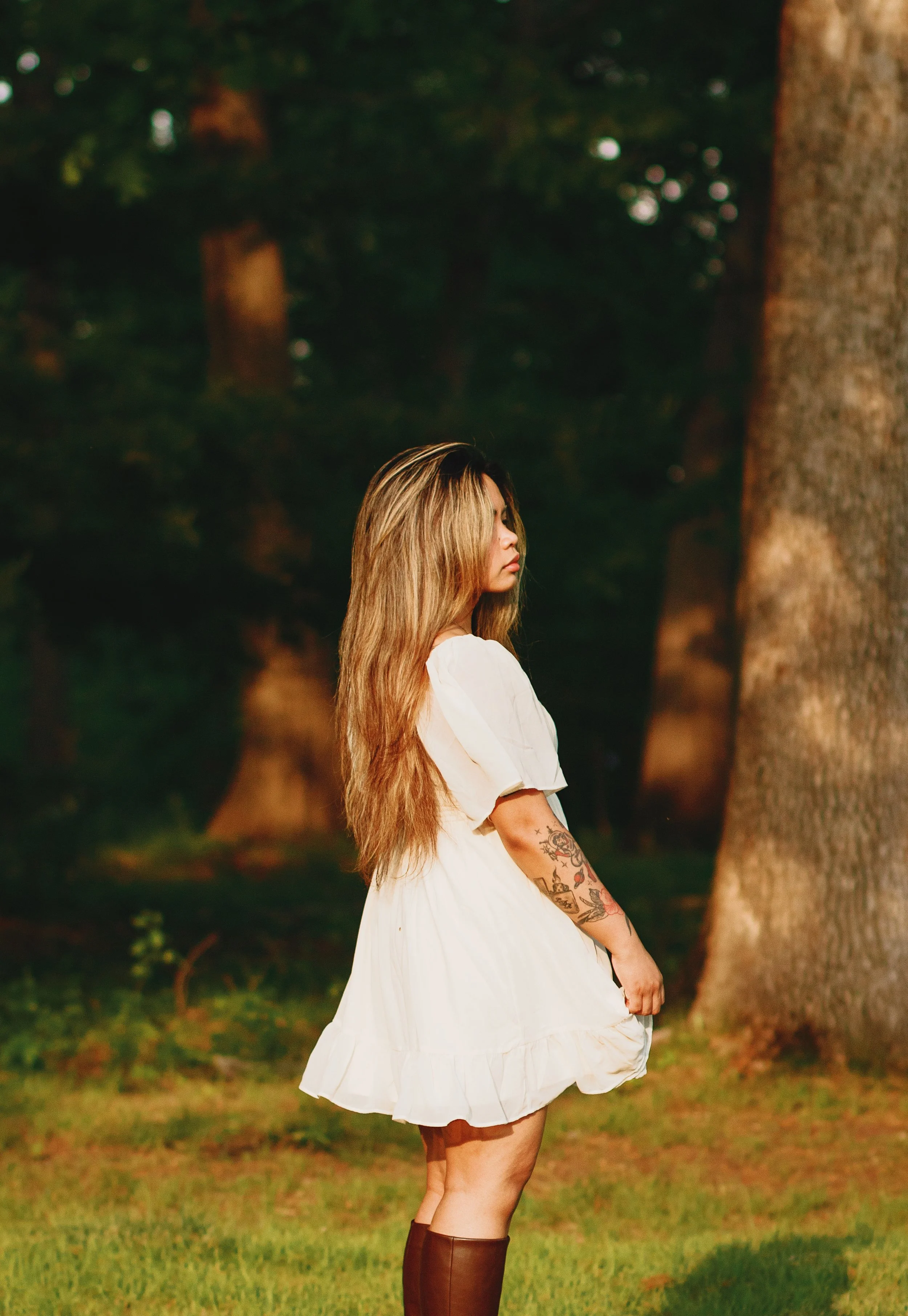 A woman with long blonde hair in a white dress standing outdoors near a tree at sunset.