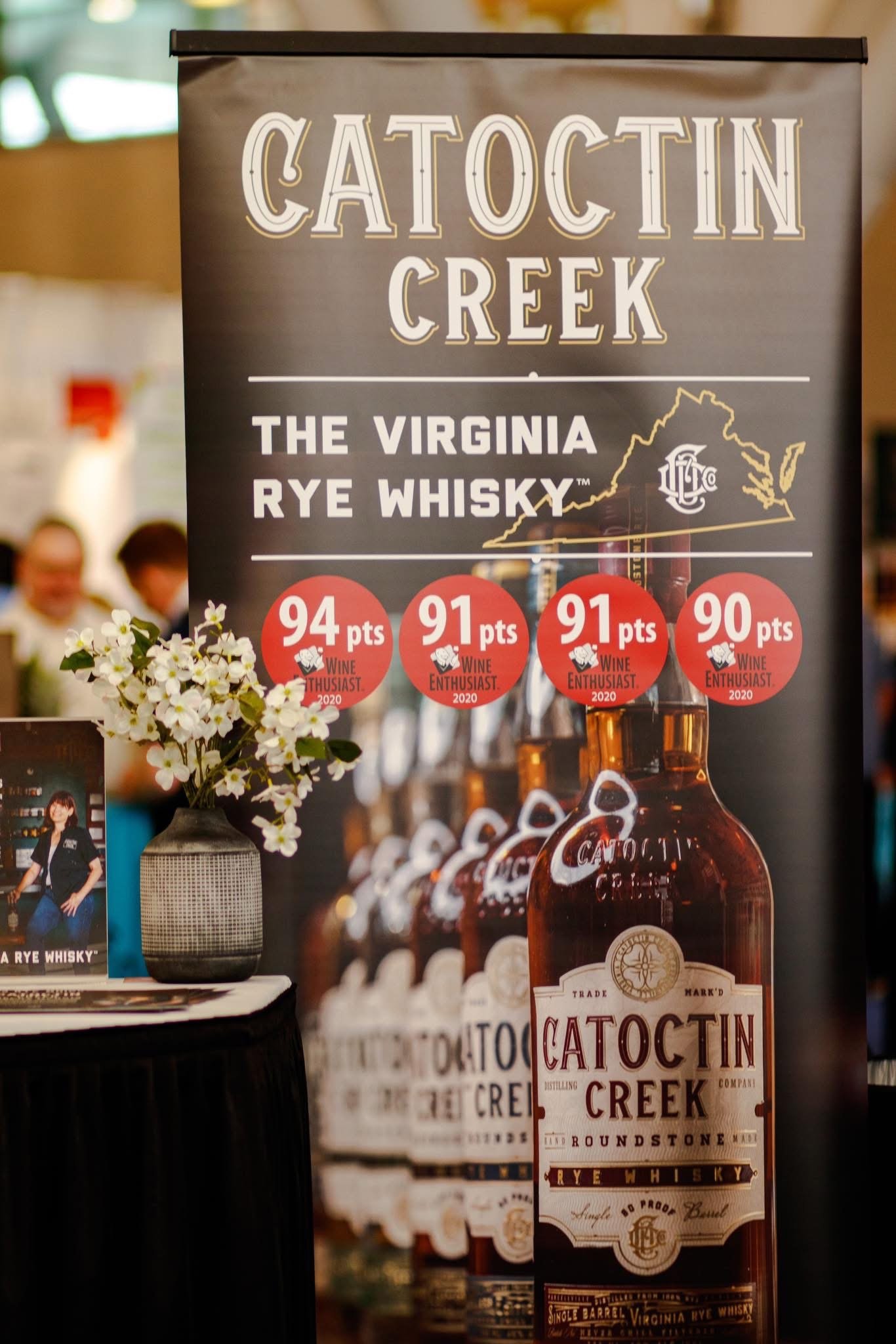 Banner advertising Catoctin Creek Virginia Rye Whisky with a map outline of Virginia and a bottle, appears to be at an event or exhibition, with people and floral arrangements in the background.