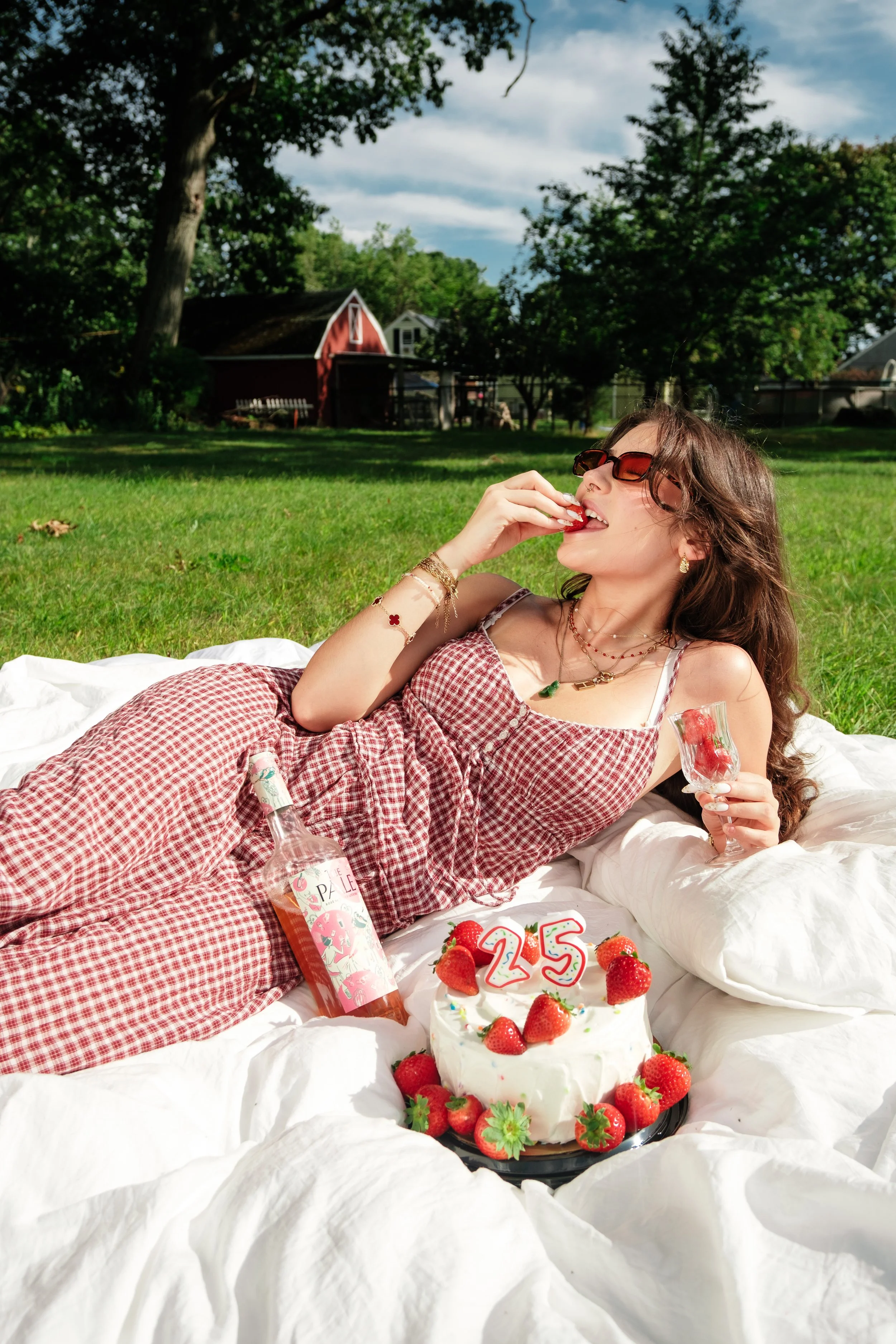 A woman lying on a white blanket outdoors, eating a strawberry, with a birthday cake and strawberries before her, celebrating her 25th birthday.