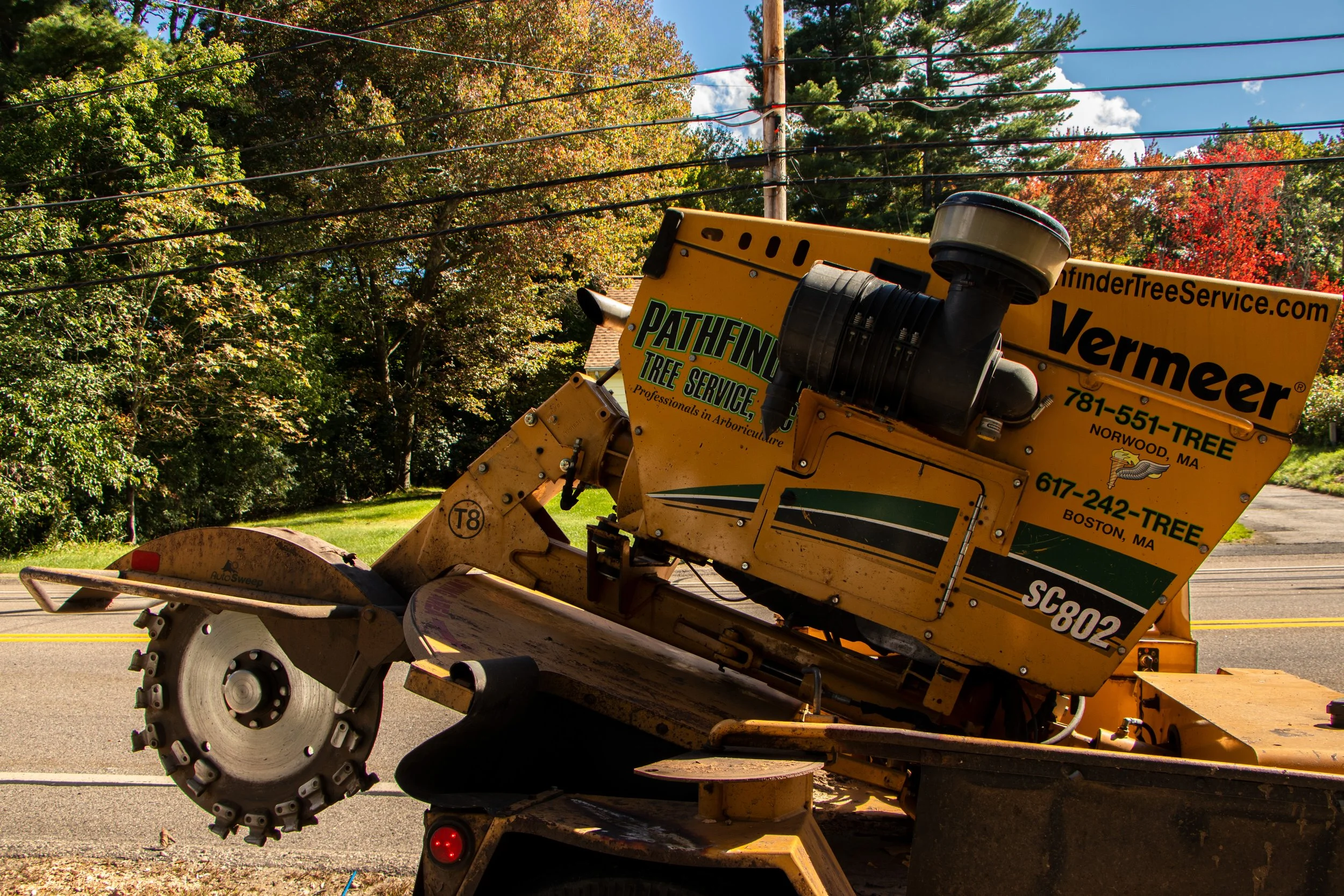 A yellow tree trimming machine on a street with trees in their fall colors and autumn leaves in the background.