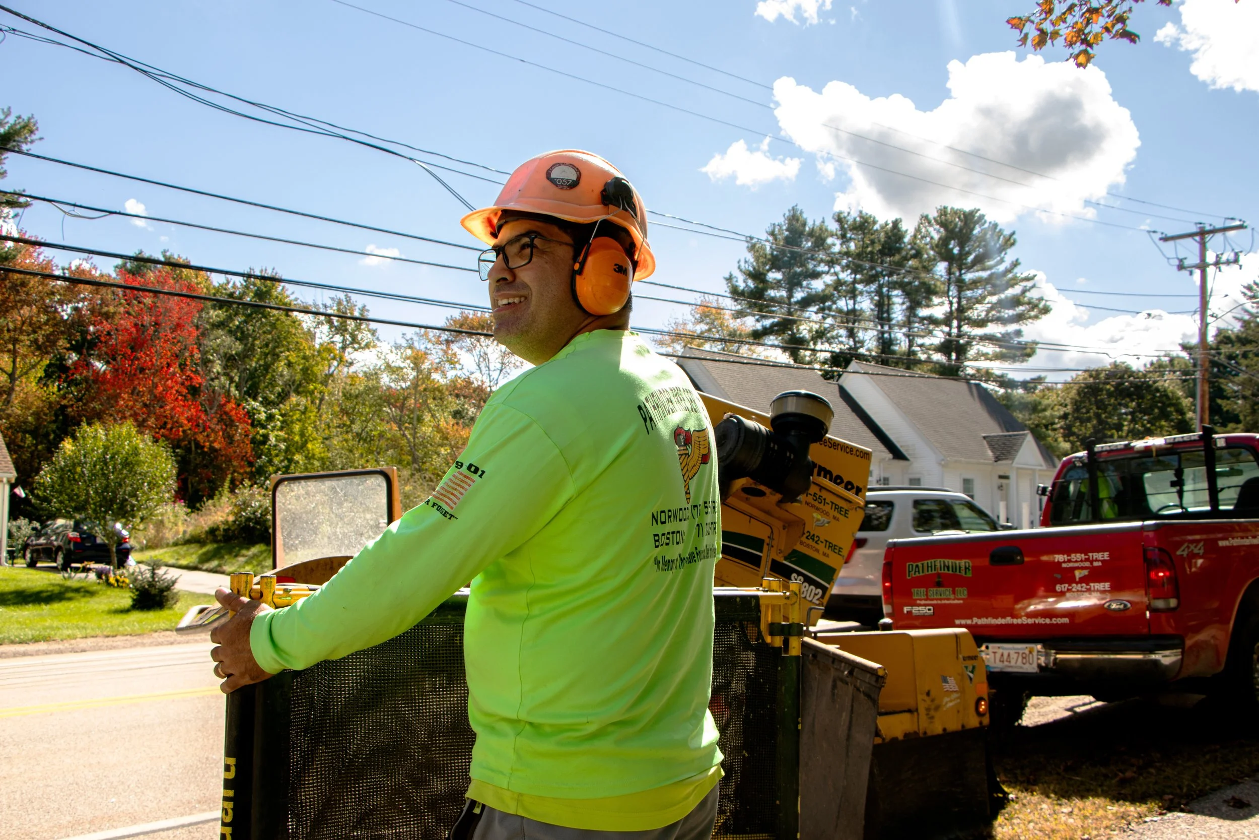 A man wearing safety glasses, a hard hat, and a high-visibility shirt is operating equipment outdoors during daytime. In the background, there are trees with fall foliage, power lines, houses, and vehicles parked along the street.