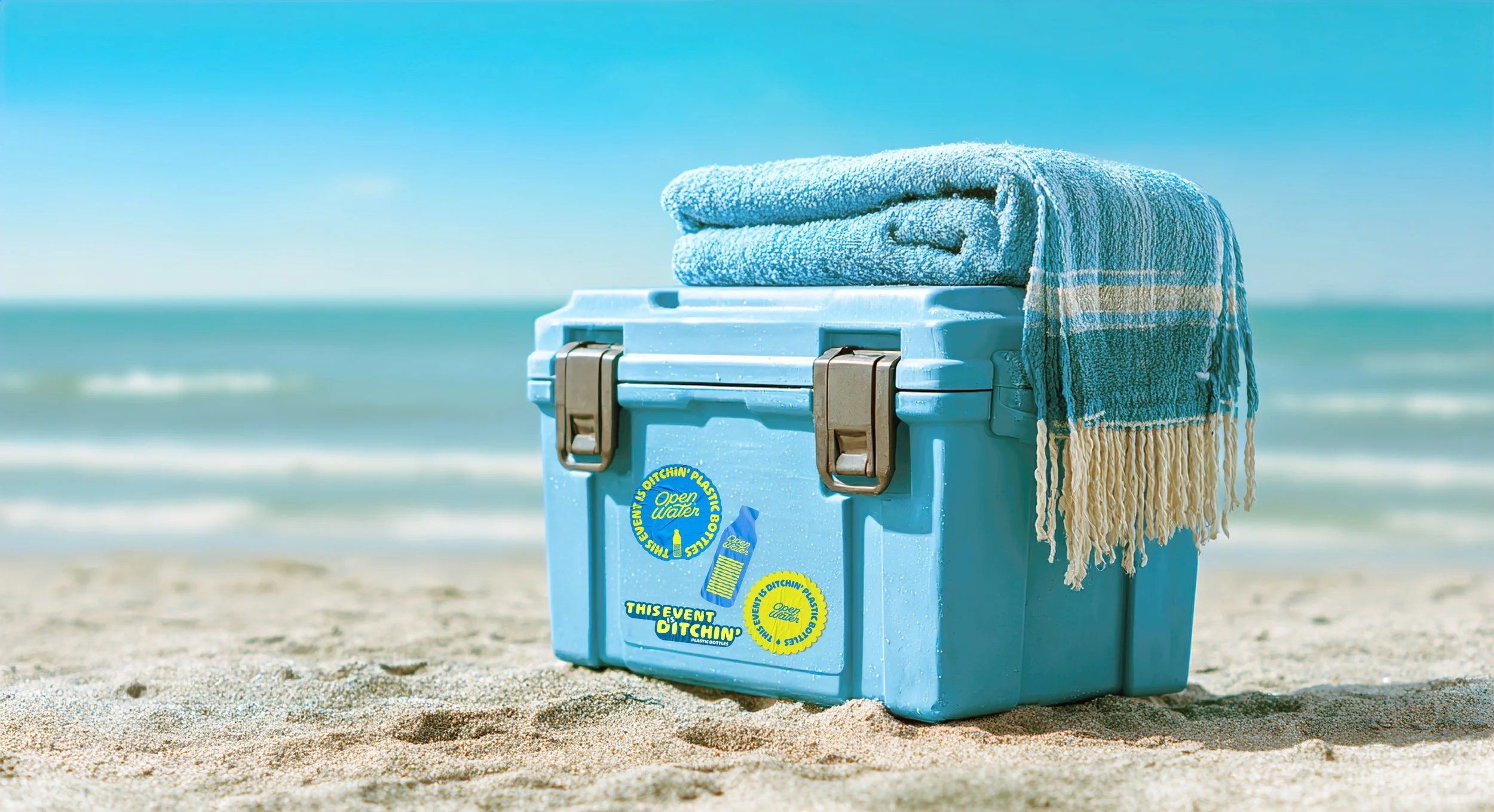 A blue plastic cooler on a sandy beach, with a rolled towel on top. The cooler has stickers that read 'This event is ditchin' plastic bottles' and 'Open water.' In the background, the ocean and sky are visible.