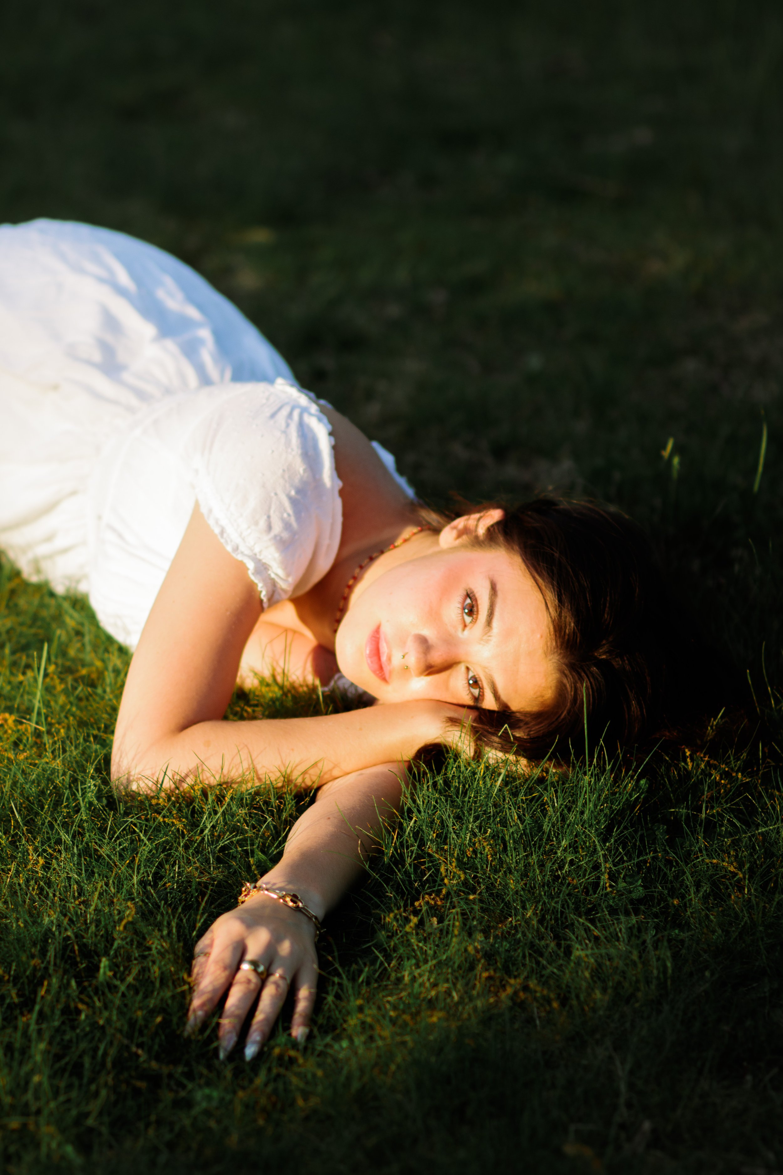 A young woman with brown hair lying on green grass in sunlight, wearing a white dress and looking at the camera.