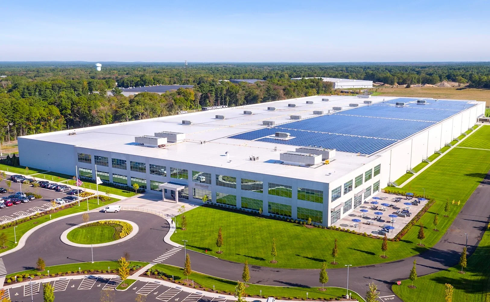 A large commercial building with a parking lot, surrounded by well-maintained green lawns and trees, with solar panels installed on the roof.
