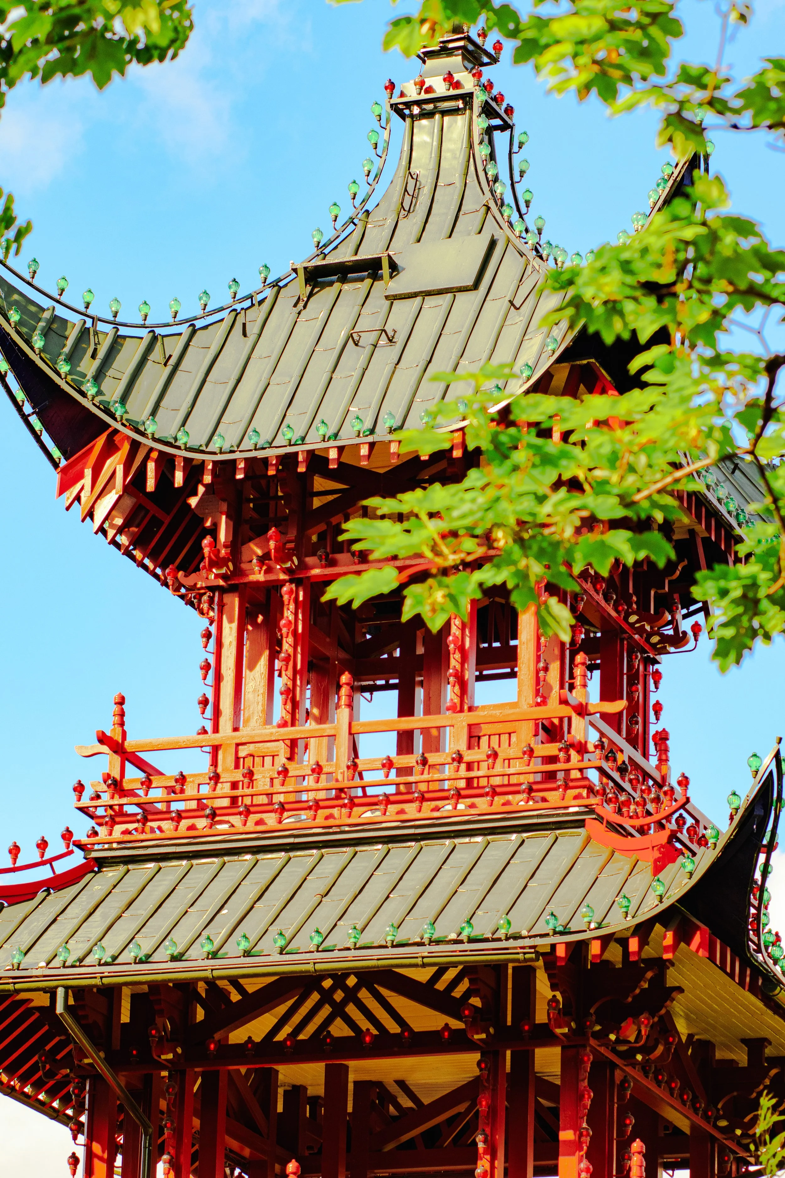 Close-up of the top section of a traditional Japanese pagoda with a green and gray tiled roof, red wooden structural elements, and decorative lighting accents, partially framed by green tree leaves against a blue sky.