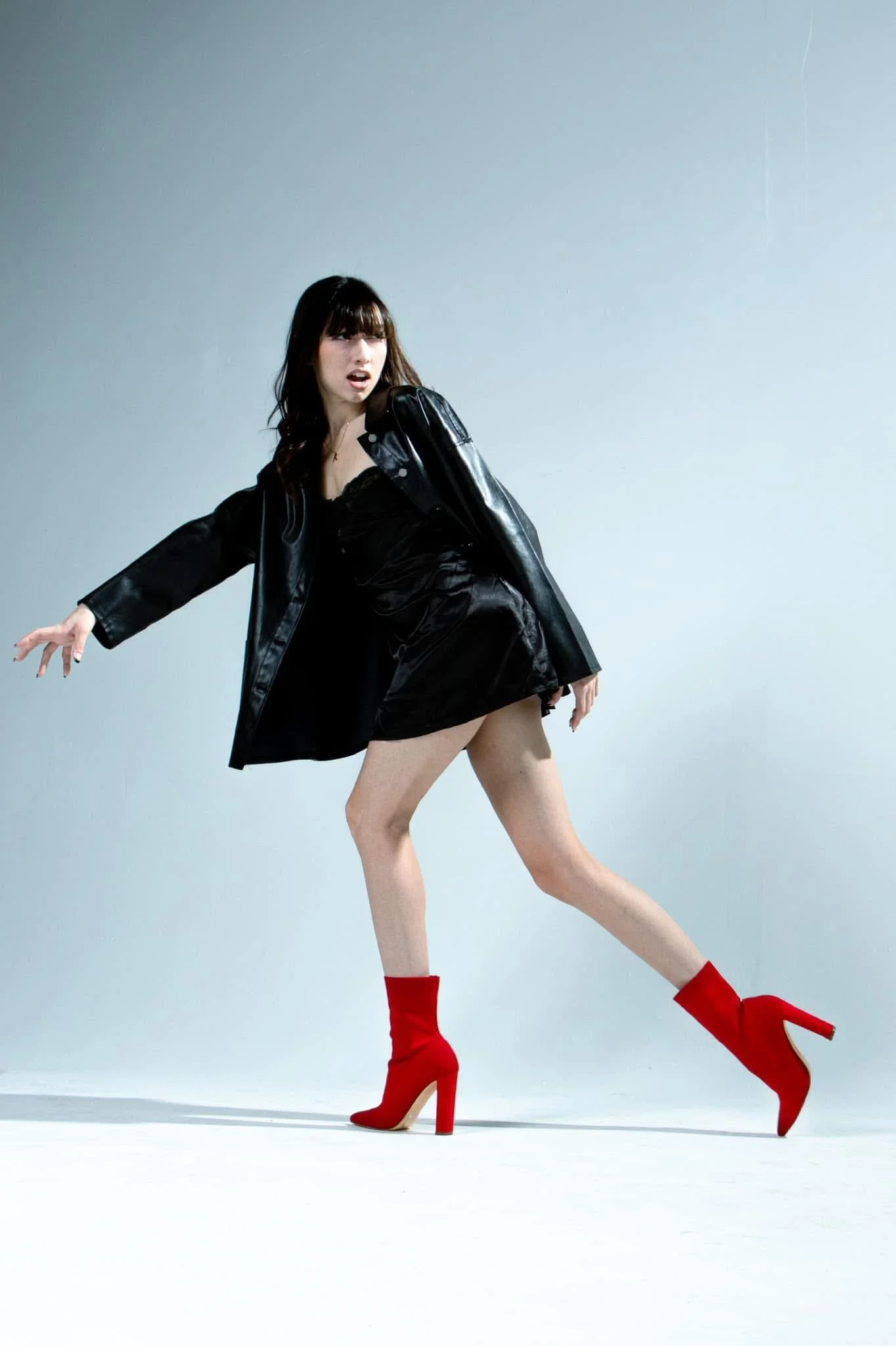 A woman wearing a black leather jacket and black dress is striking a pose against a plain light gray background. She is wearing bright red high-heeled boots.