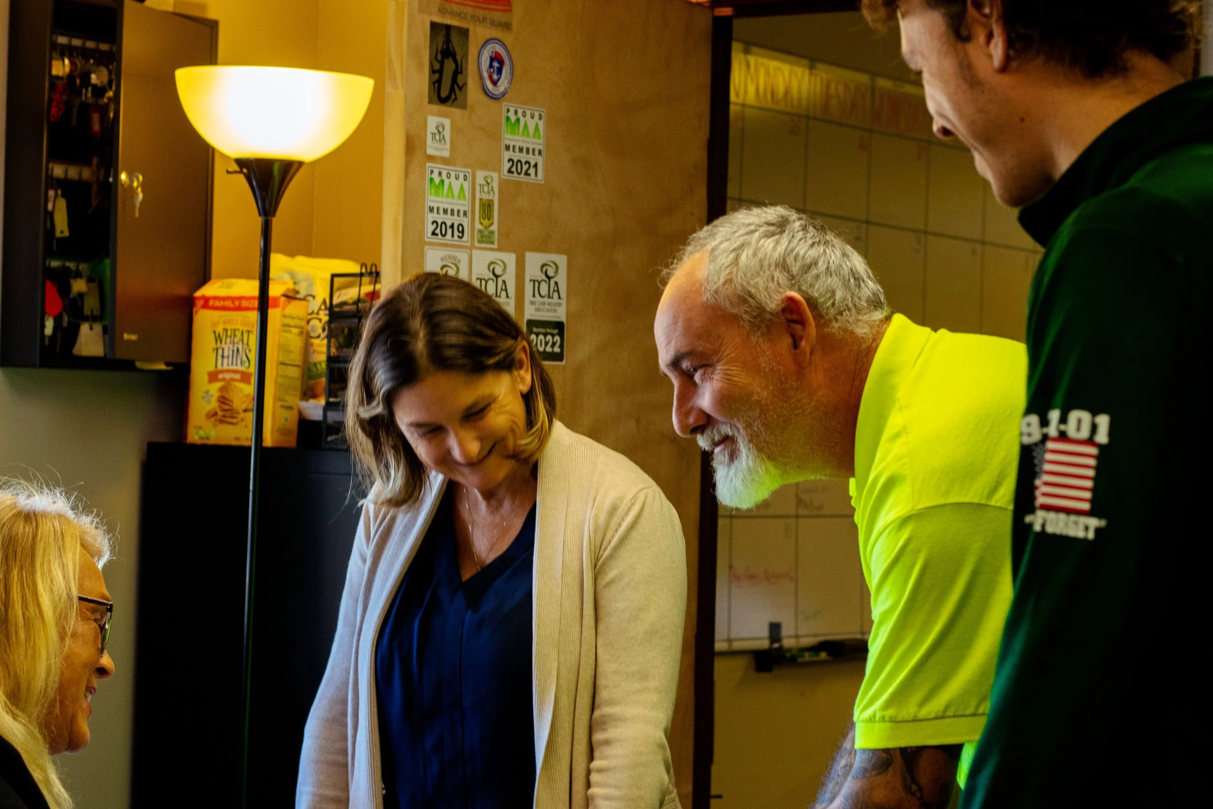 A group of four people are gathered indoors, engaged in conversation and smiling. One woman with glasses and blonde hair is sitting, while a woman with shoulder-length brown hair is standing next to her. Behind them, a man with gray hair and a beard,