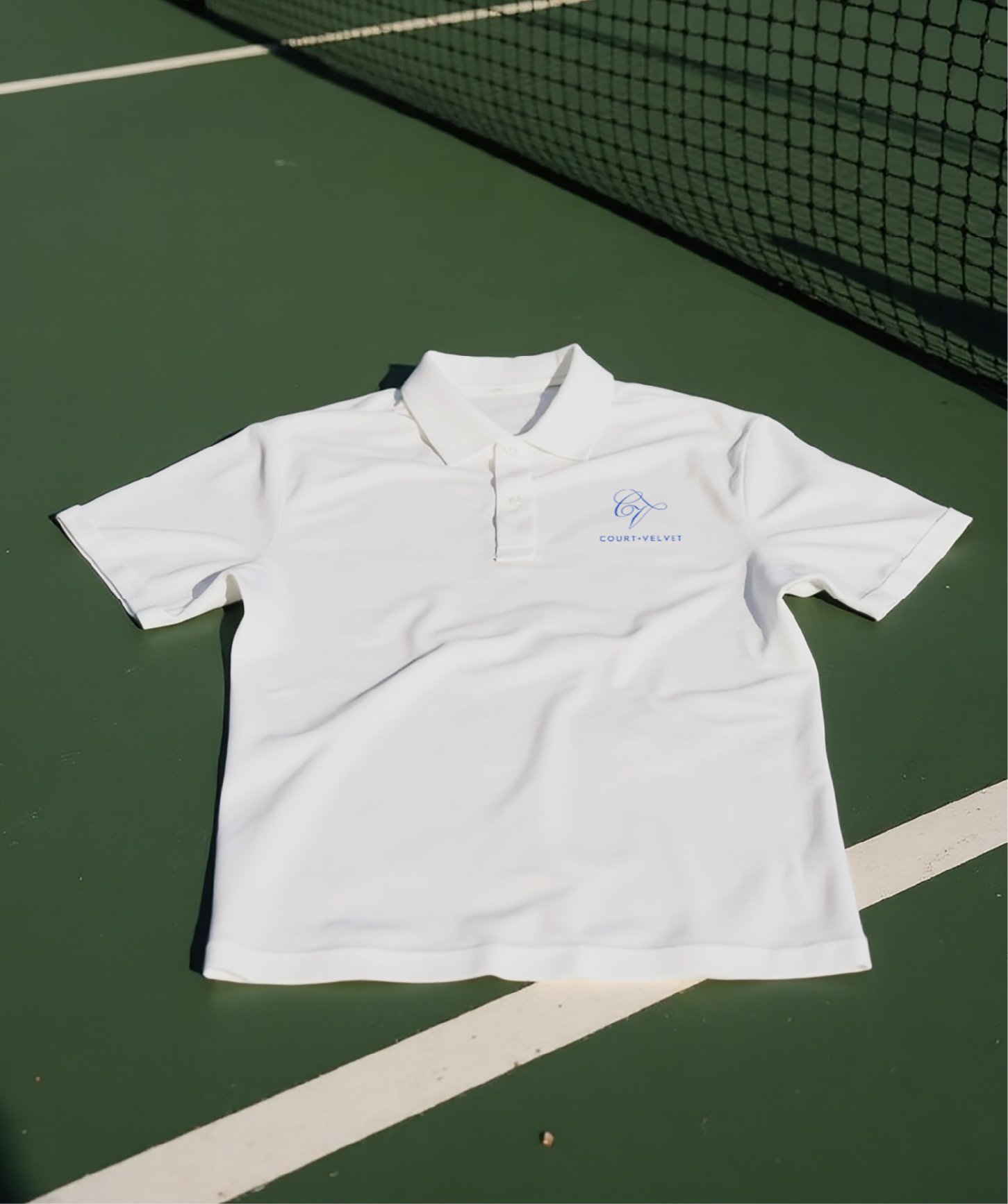 White polo shirt with blue CV logo and text lying on a green tennis court near the net.