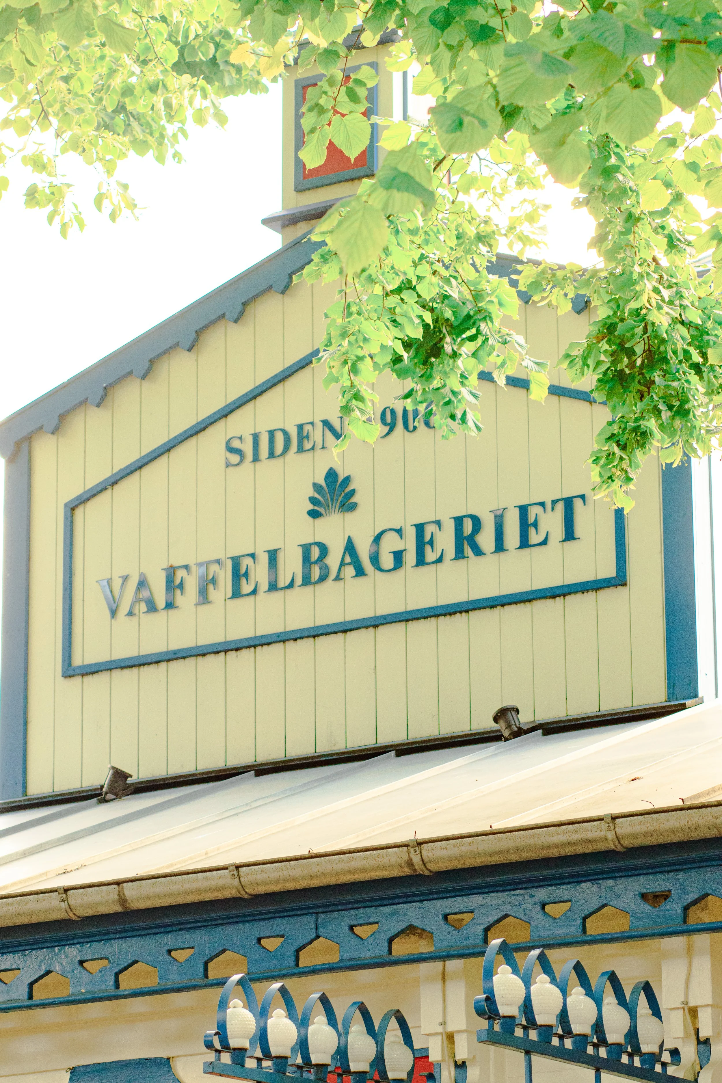 Close-up of a yellow building with a sign reading 'Vaffelbakeren' and part of a roof with a gutter, surrounded by green leaves from a nearby tree.