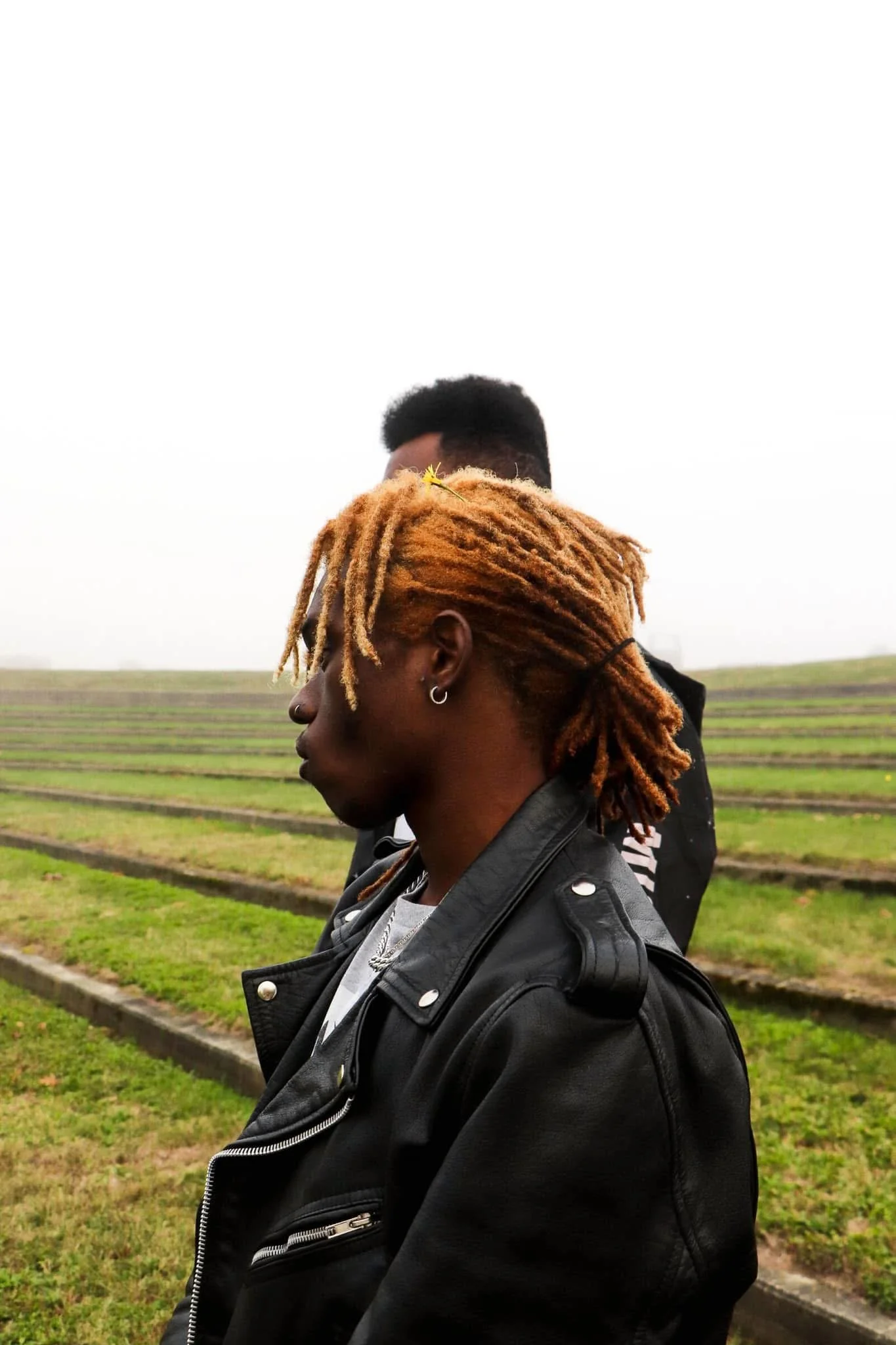 Profile of a person with tan dreadlocks and piercings, wearing a black leather jacket, standing outdoors with fields in the background.