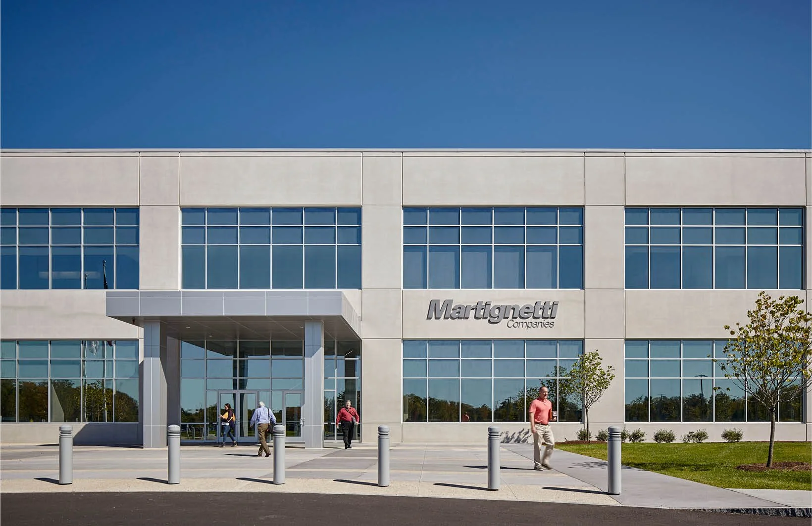 Front view of a modern office building with a sign that reads 'Martignetti Companies'. Several people are walking near the entrance, which is surrounded by small trees and a neatly maintained lawn; bollards line the driveway, and the sky is clear and blue.