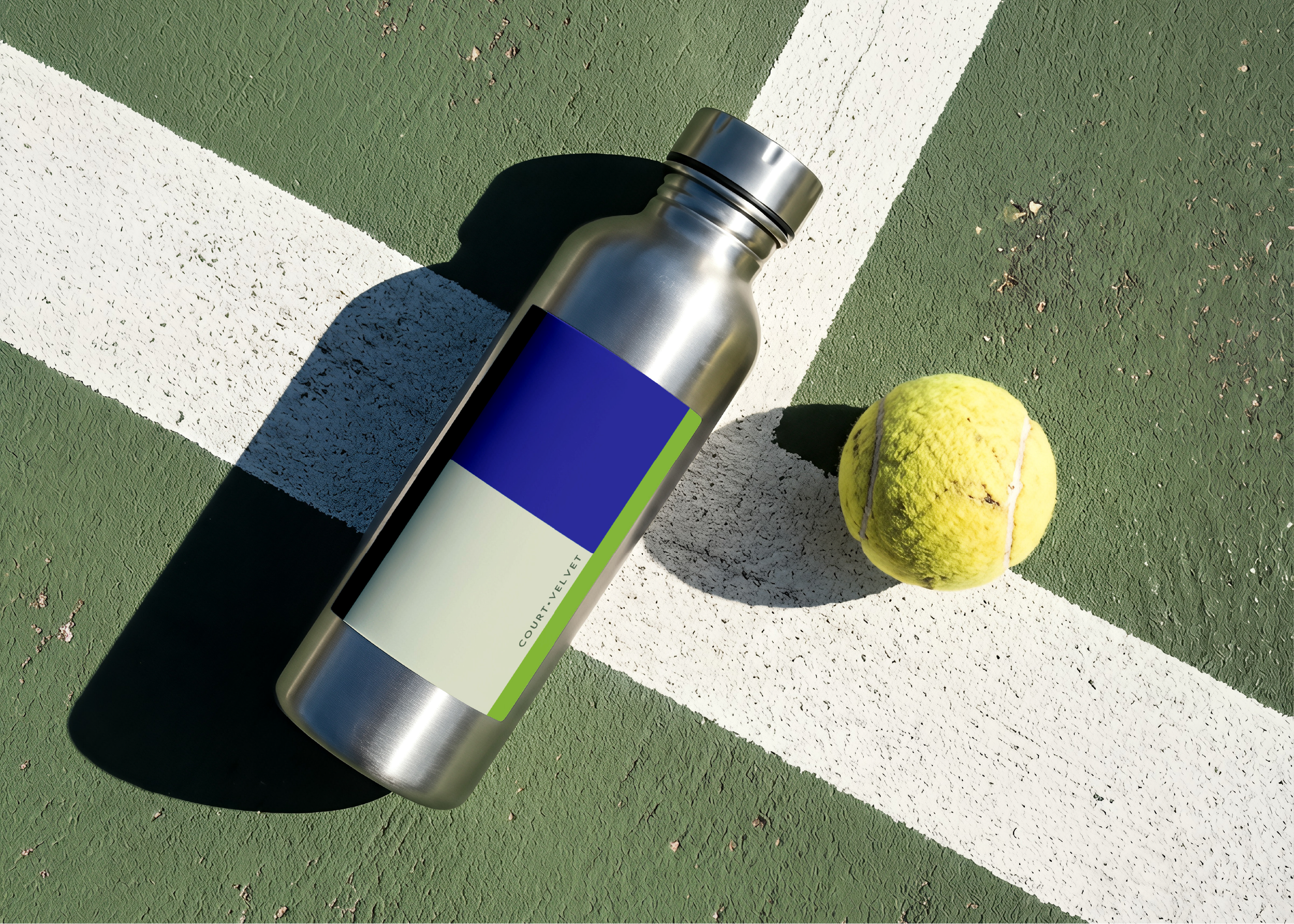 A stainless steel water bottle with a blank blue and green label lying on a green and white textured tennis court next to a yellow tennis ball.