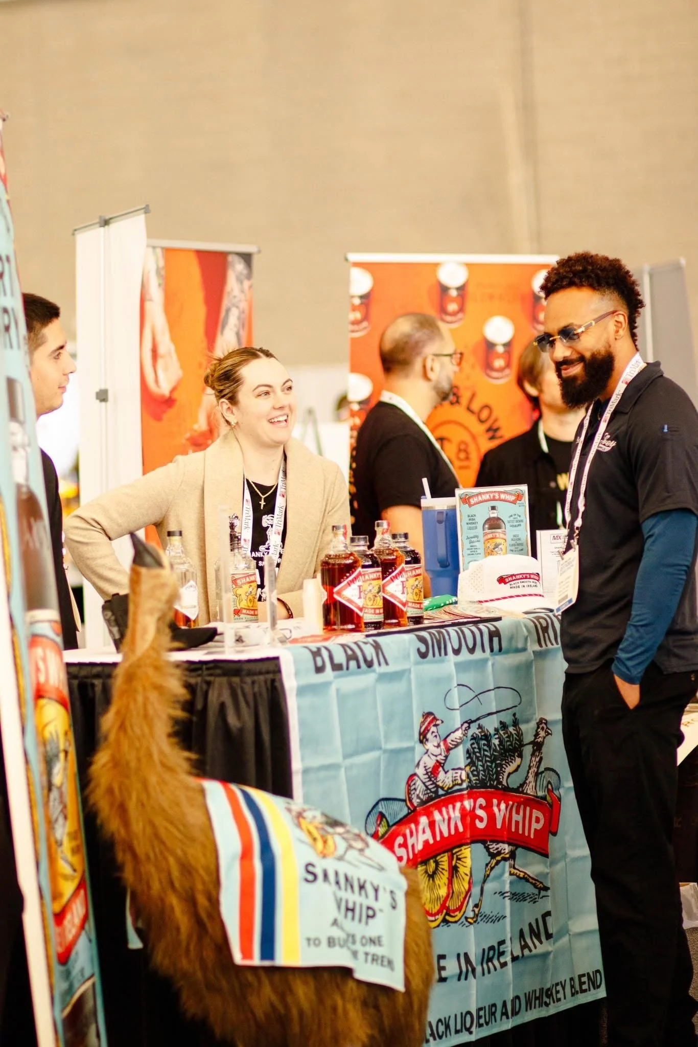 People attending a booth at an event, with various product bottles on display, and a large colorful banner advertising Shanky's Whip. A person in the foreground with a brown fur coat and a banner on its back.