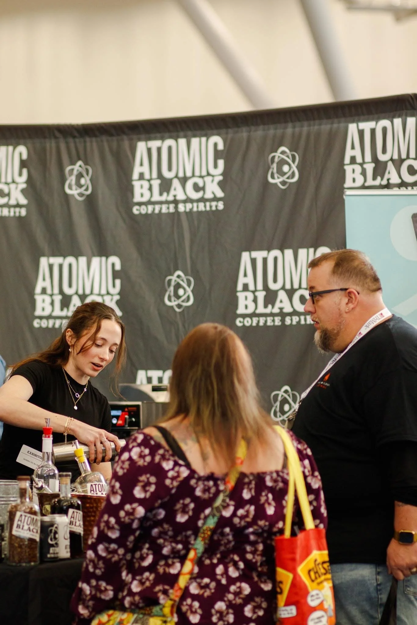 People at a booth or display for Atomic Black Coffee Spirits, with various bottles and products on the table.