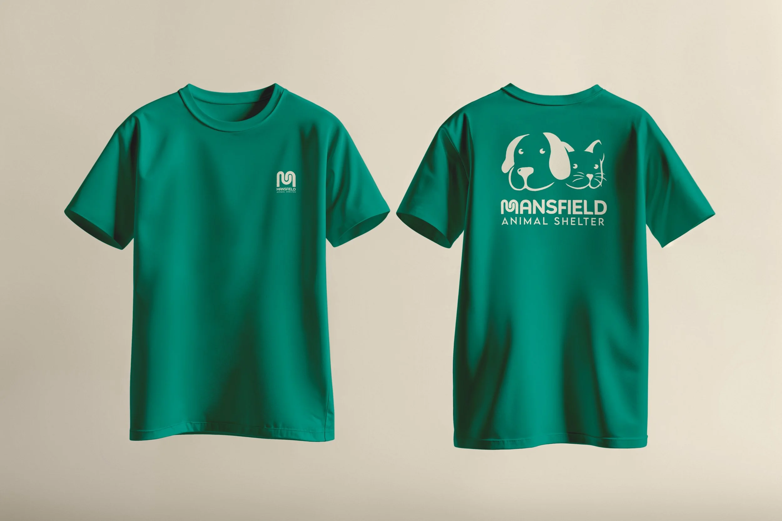 Two teal T-shirts hanging side by side on a light background. The front of the left shirt has a small logo reading 'MANSFIELD Animal Shelter'. The back of the right shirt features a large graphic of a dog's and a cat's face, along with the text 'MANSFIELD Animal Shelter'.