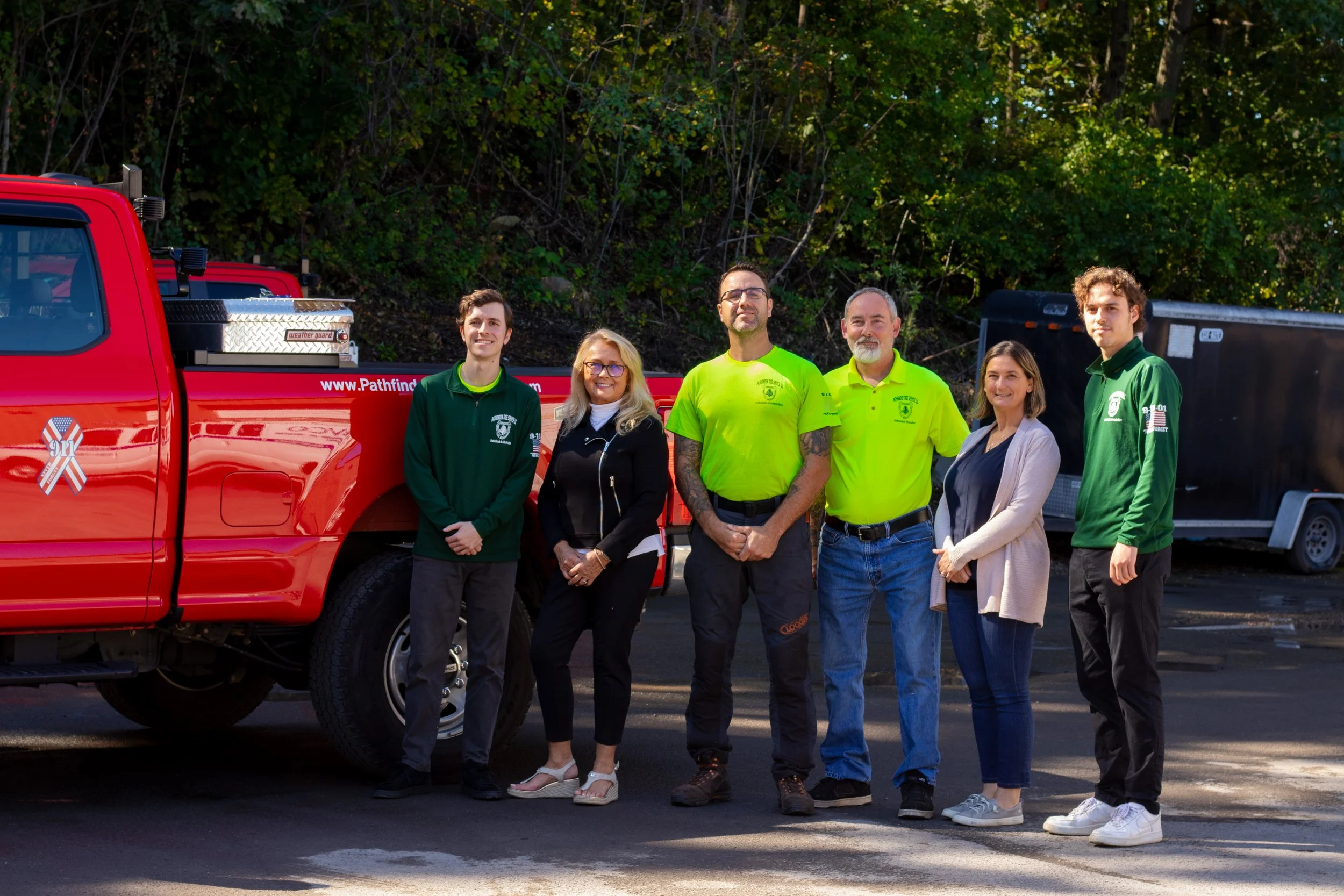 A group of six people standing outdoors in front of a red emergency response vehicle with trees in the background. The group includes women and men, some wearing official-looking clothing, smiling and posing for the photo.