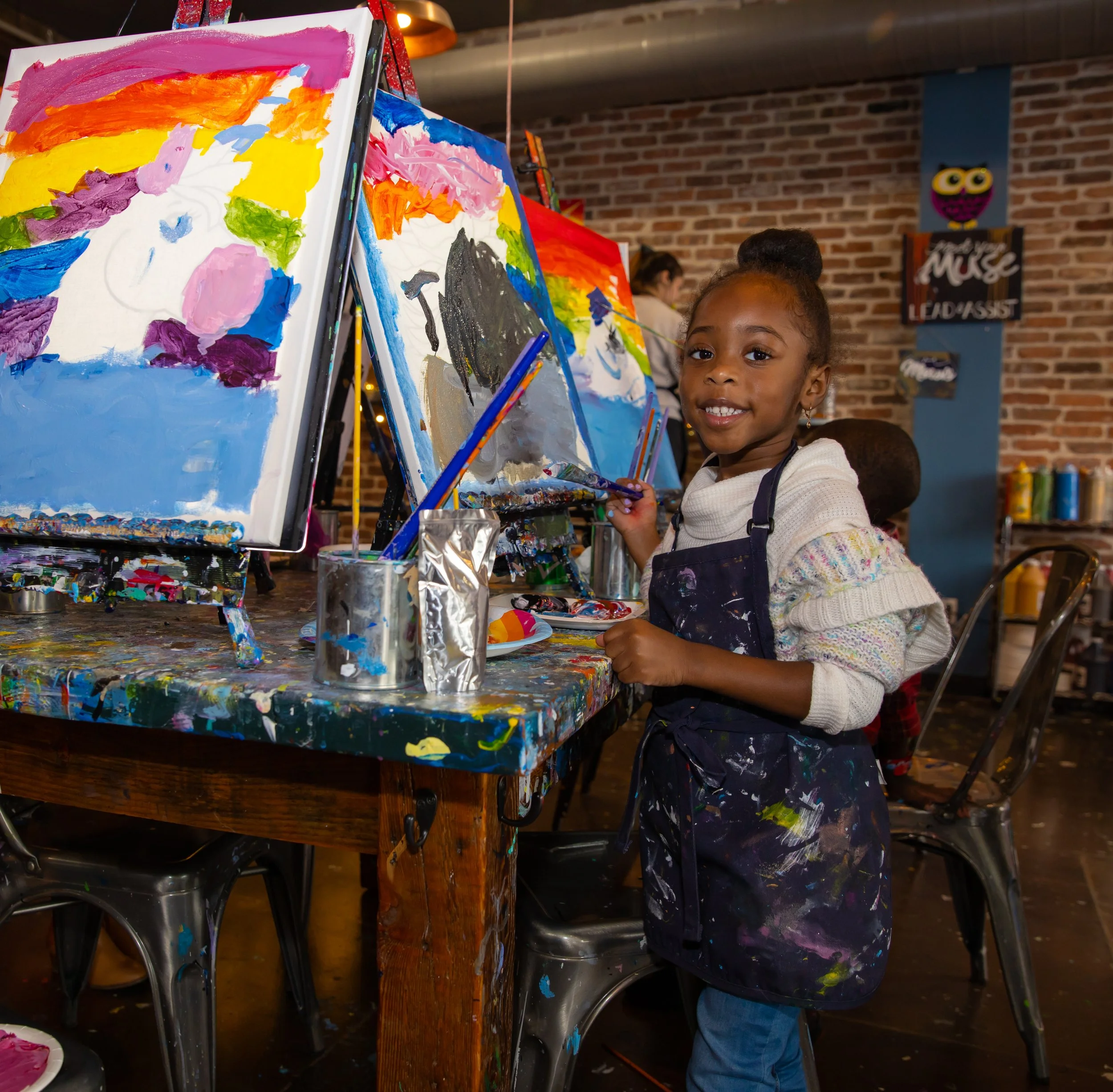 A young girl smiling as she paints on a canvas at an art event. The canvas features bright colors and abstract shapes. The background shows a brick wall and other children engaged in painting.