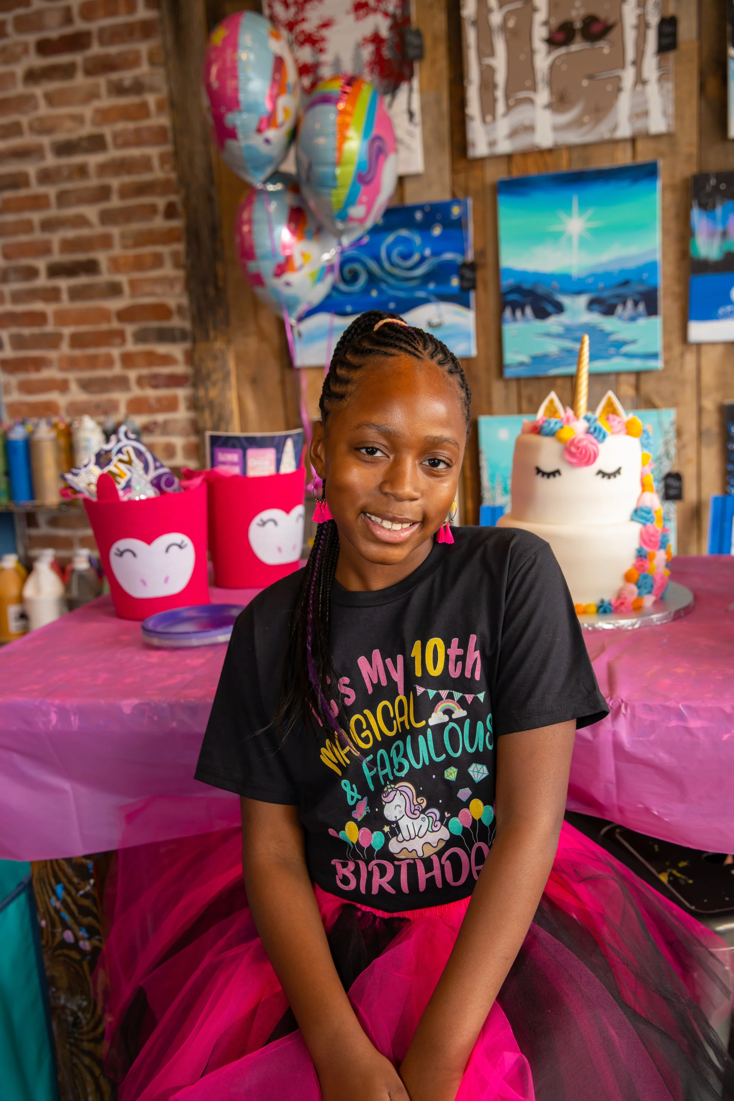 A young girl at her birthday party, wearing a black T-shirt with colorful text, a pink tutu, and pink earrings, standing in front of a decorated table with a unicorn-themed birthday cake and balloons in the background.