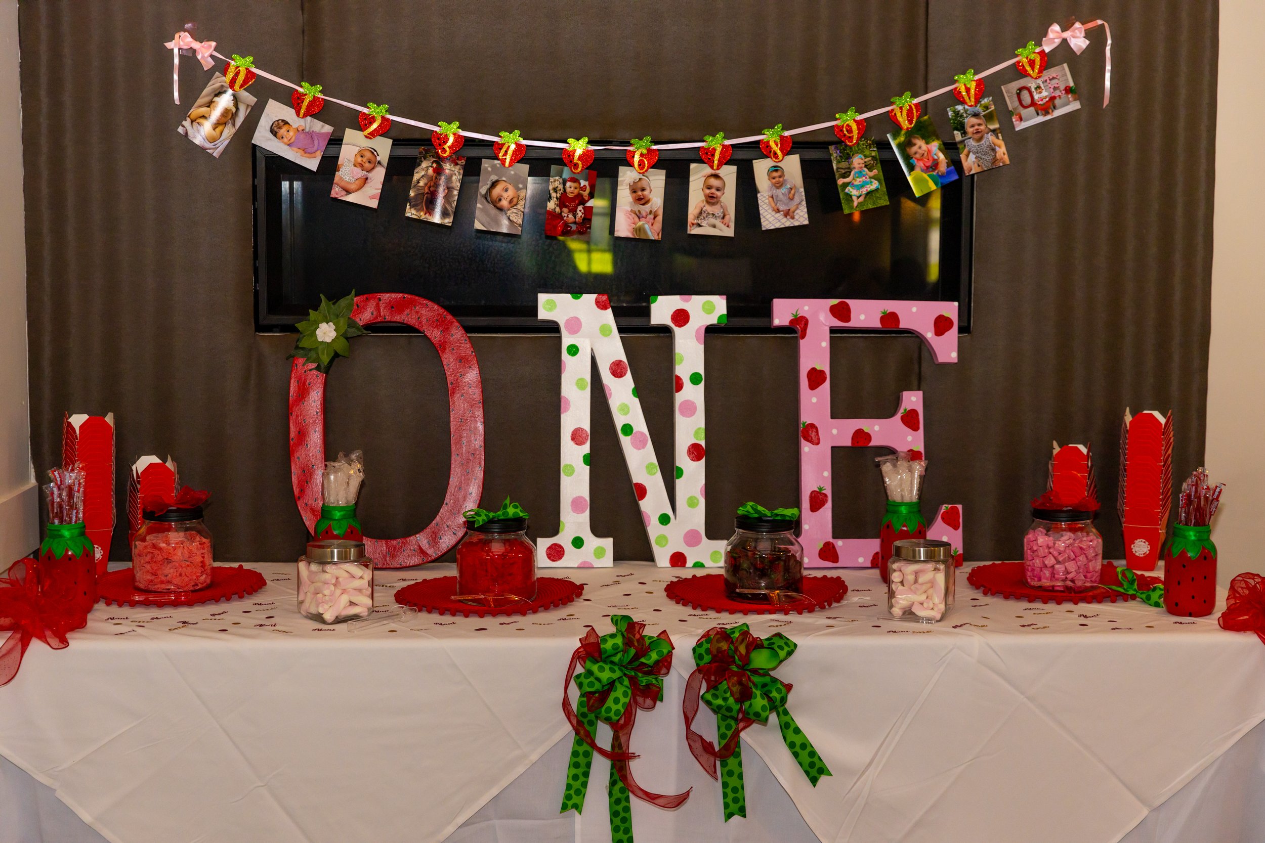 Table decorated for Valentine's Day with large colorful letters spelling 'LOVE', jars of candies, and a Valentine-themed banner featuring photos of children, strawberries, and presents.