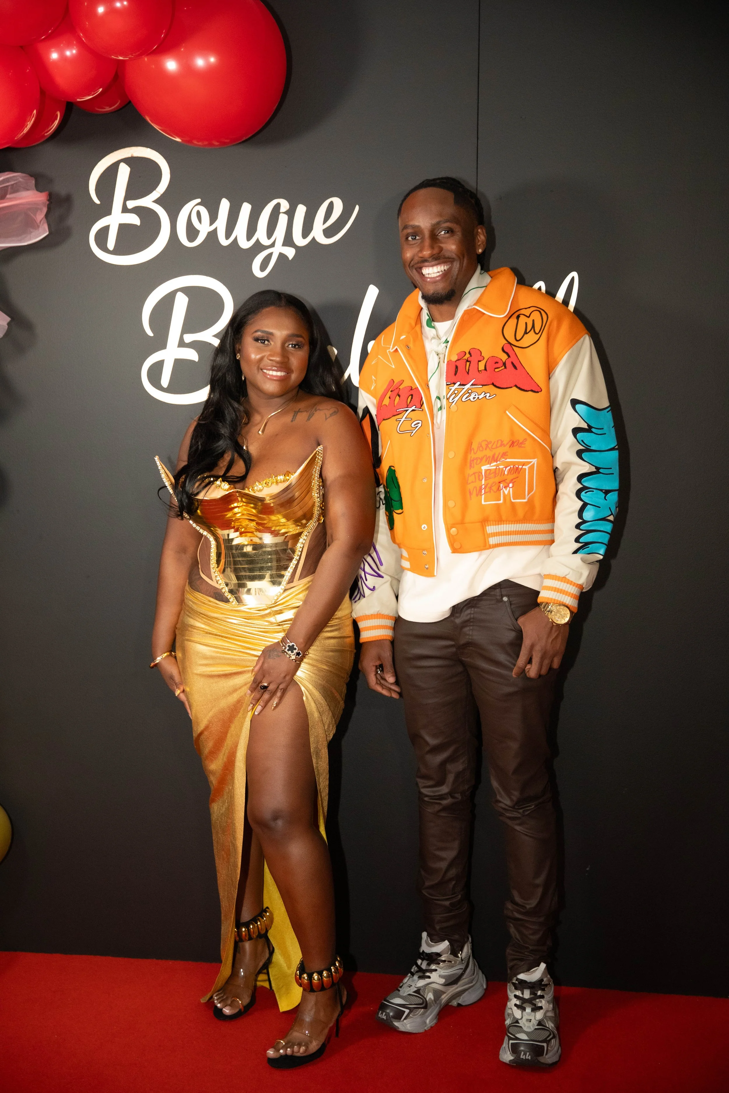 A woman in a gold dress with a high slit, black high heels, and jewelry, standing next to a man in casual sporty clothing with a bright jacket, on a red carpet at an event with a black background that has white text and large red balloons.