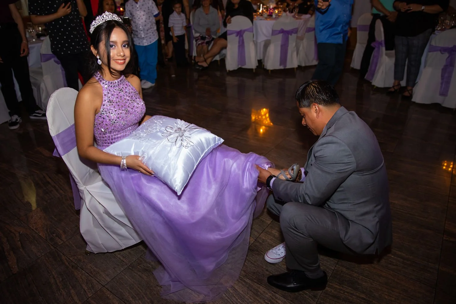 A girl in a purple dress and tiara sitting on a chair at a celebration event, while a man in a gray suit kneels in front of her, removing her shoe. The room has a dark wood floor and tables with purple ribbons and lit candles in the background, with 