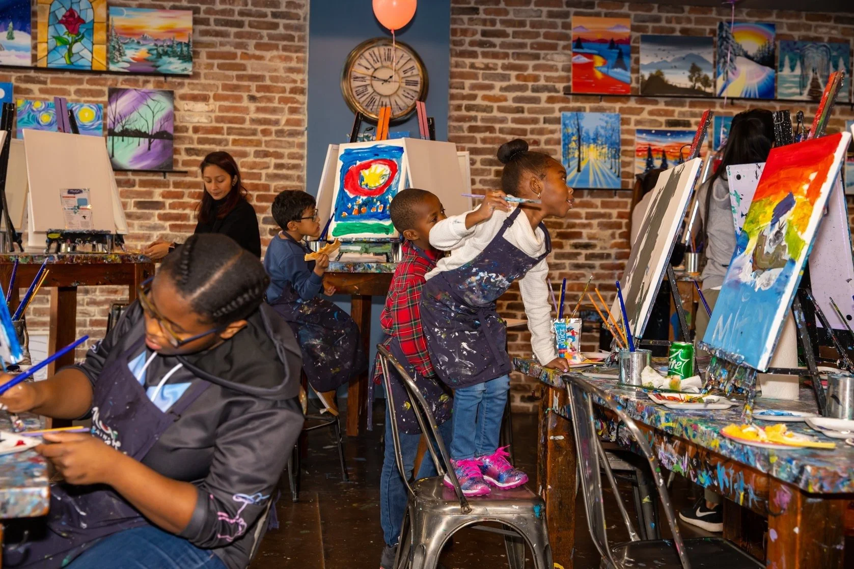 Children and adults painting on canvases in a colorful art studio with brick walls, artwork, and supplies.