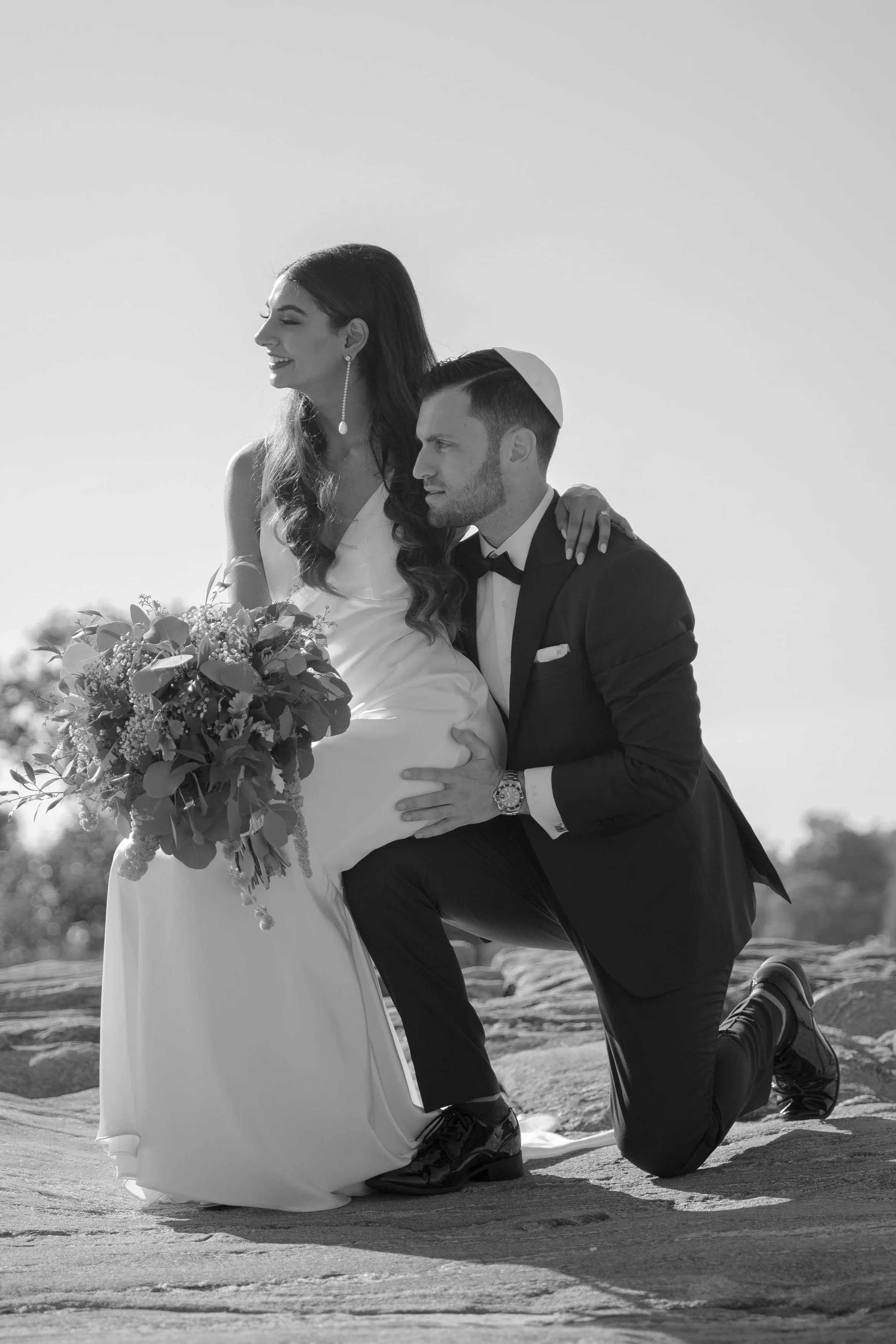 A groom kneeling on one knee, holding the bride's waist, with the bride holding a bouquet and smiling, outdoors under a clear sky.