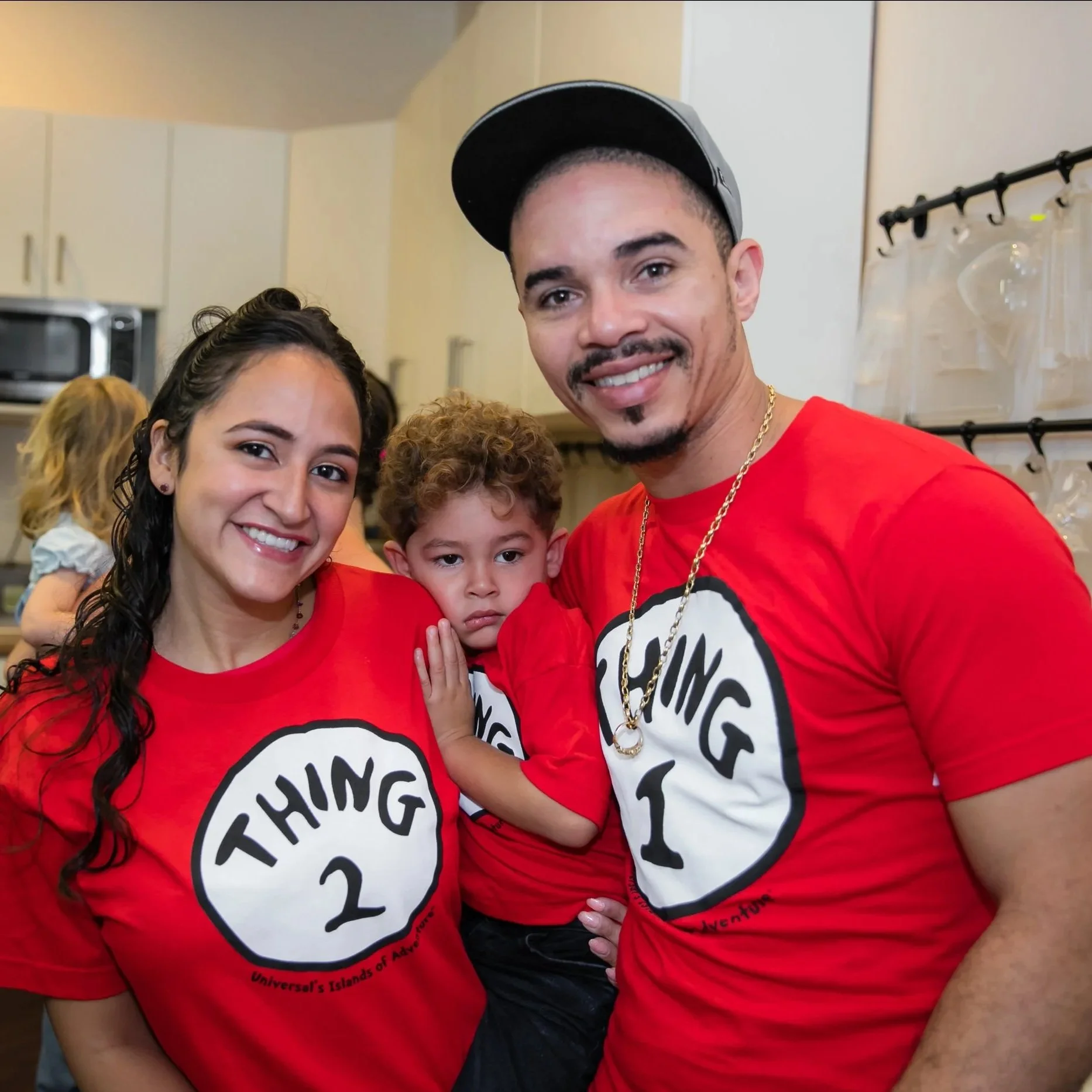 Family celebrating at a party, wearing matching red T-shirts with a white speech bubble design and black text, in a kitchen with cabinets and a microwave in the background.