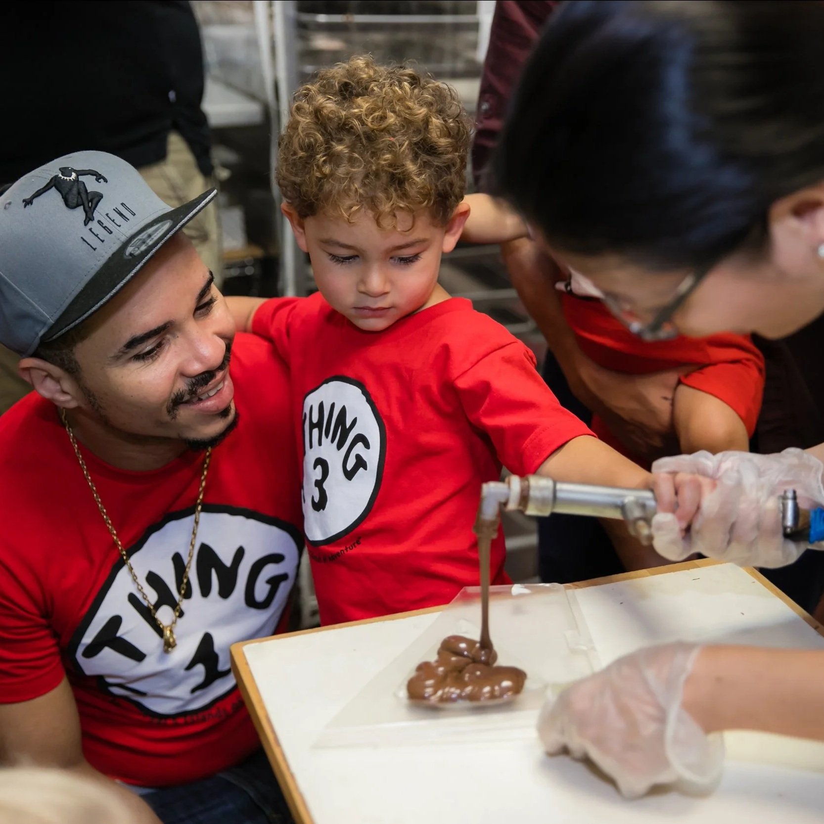 A young boy with curly hair is focusing on a science experiment involving chocolate. Two adults are assisting him at a table covered with a white tray and gloves, with a few objects and equipment around.