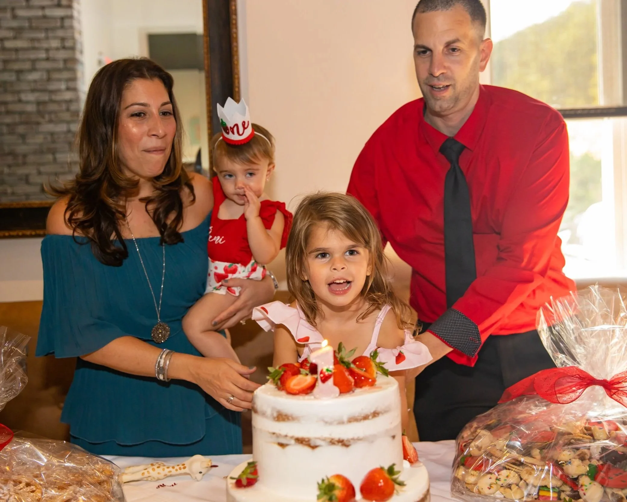 A family celebrating a birthday with a cake decorated with strawberries and a small lit candle, surrounded by party decorations and gifts.