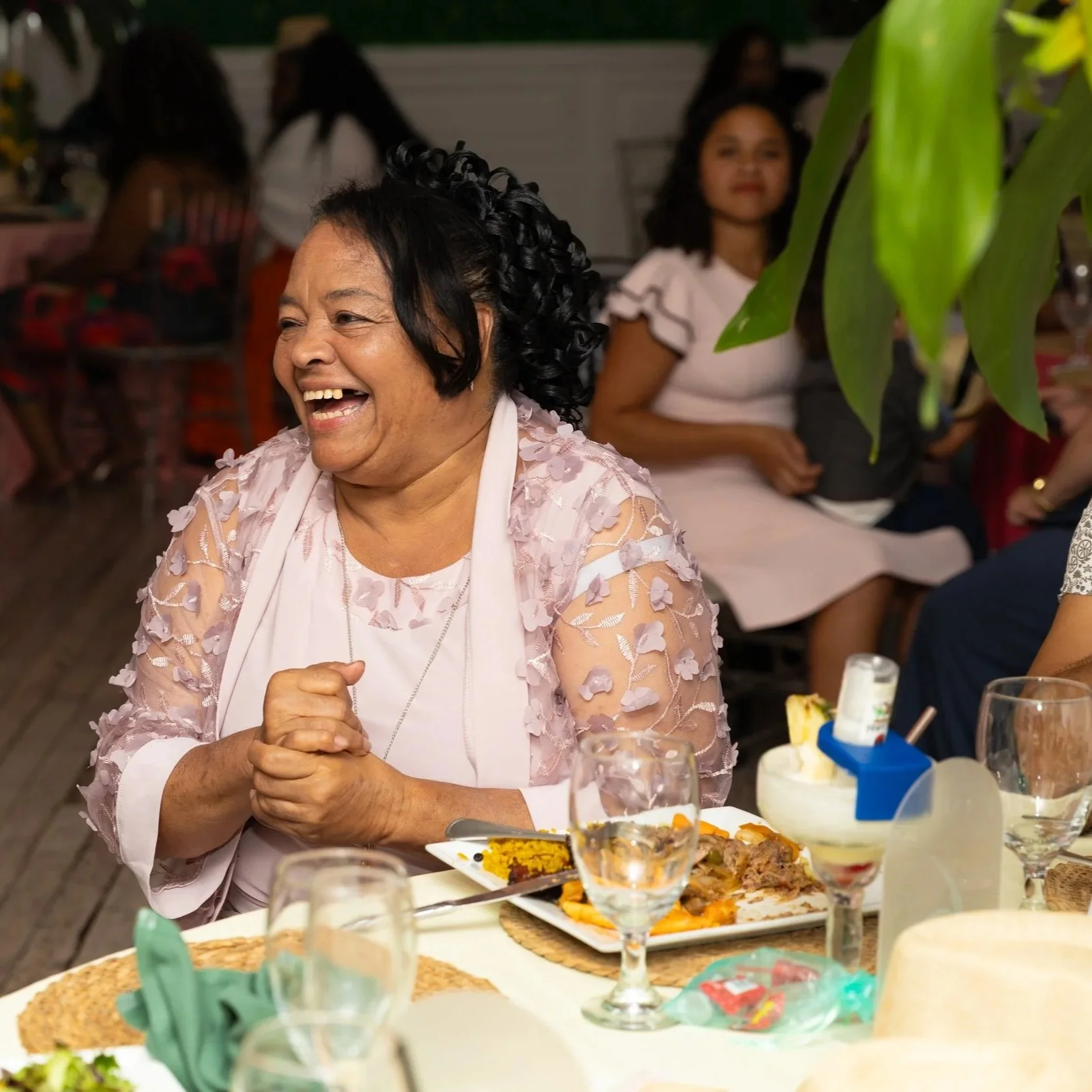 An elderly woman with black curly hair laughing while seated at a dinner table with food, glasses, and decorations, during a celebration with friends or family.