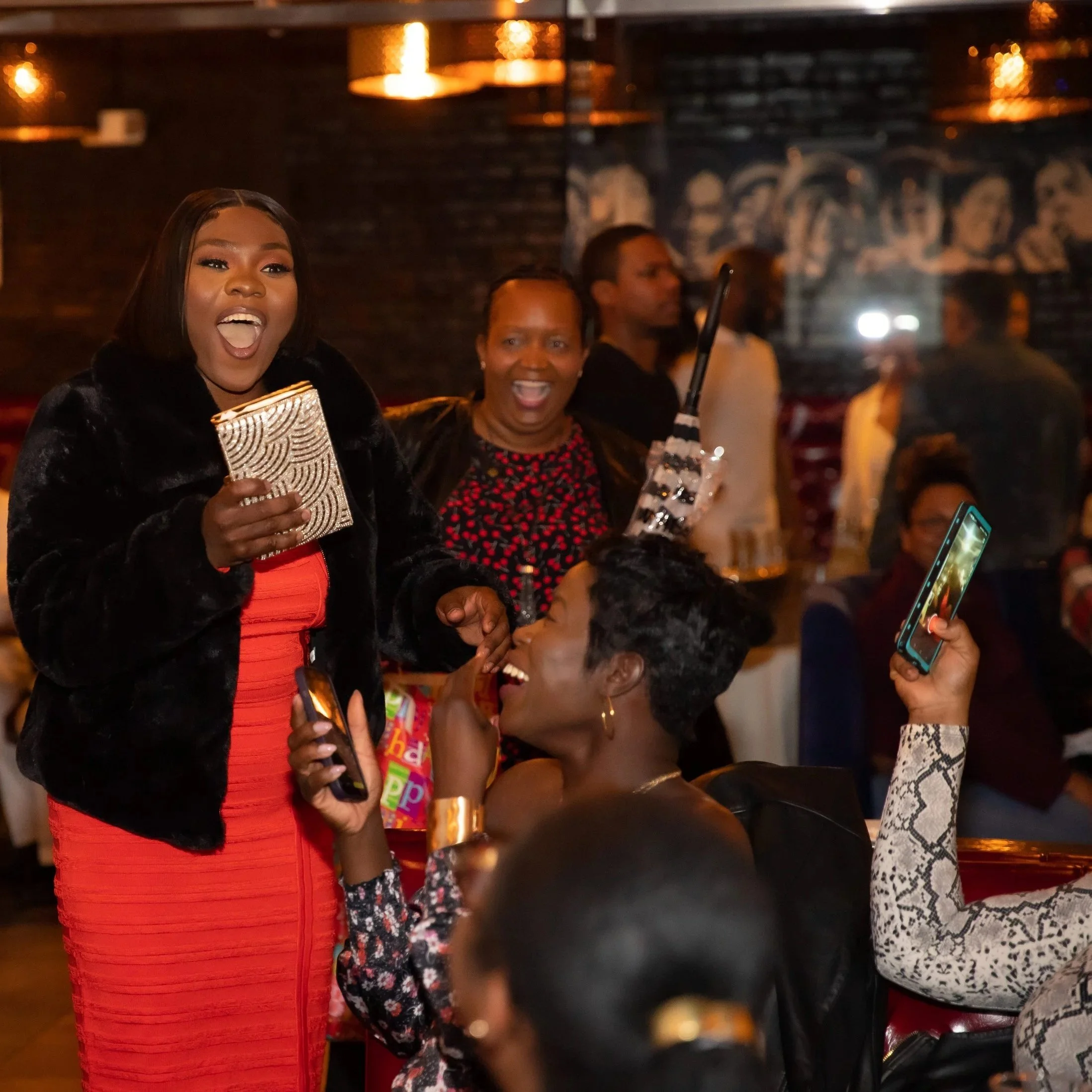 Four women are gathered together at a social event, smiling and enjoying themselves. One woman in the foreground is holding a gold clutch purse and some kind of gift, while others are smiling and holding drinks or phones. The background features a da