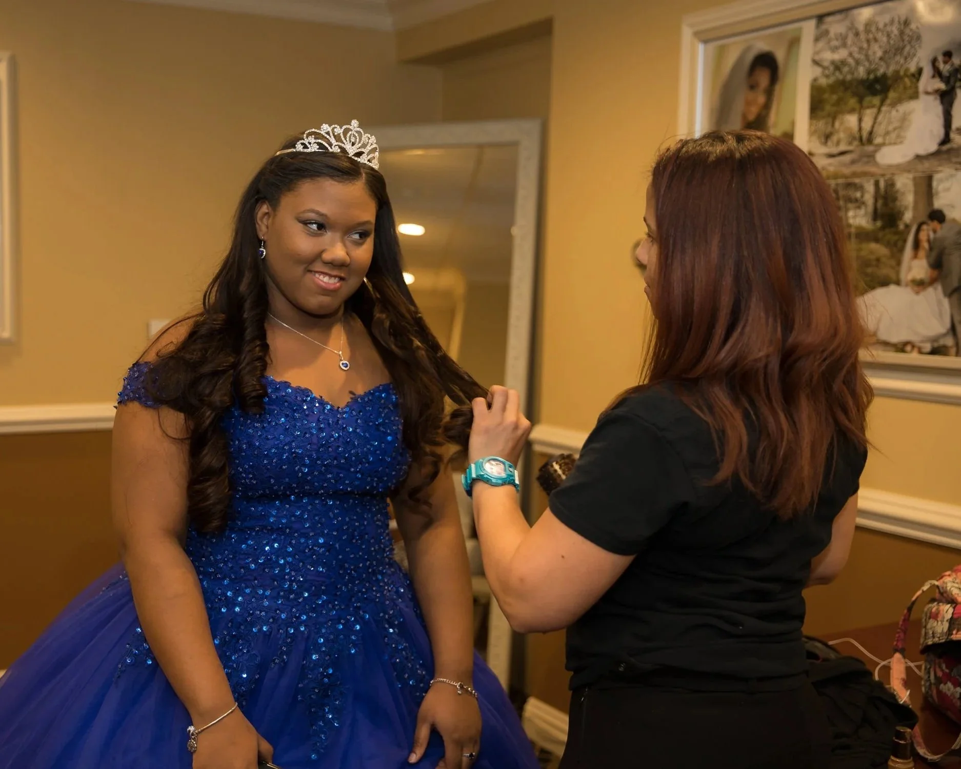 A young woman in a royal blue, sequined gown and a tiara getting her hair done by a stylist. The stylist is a woman with reddish hair, wearing a black shirt, and is adjusting the young woman's hair.