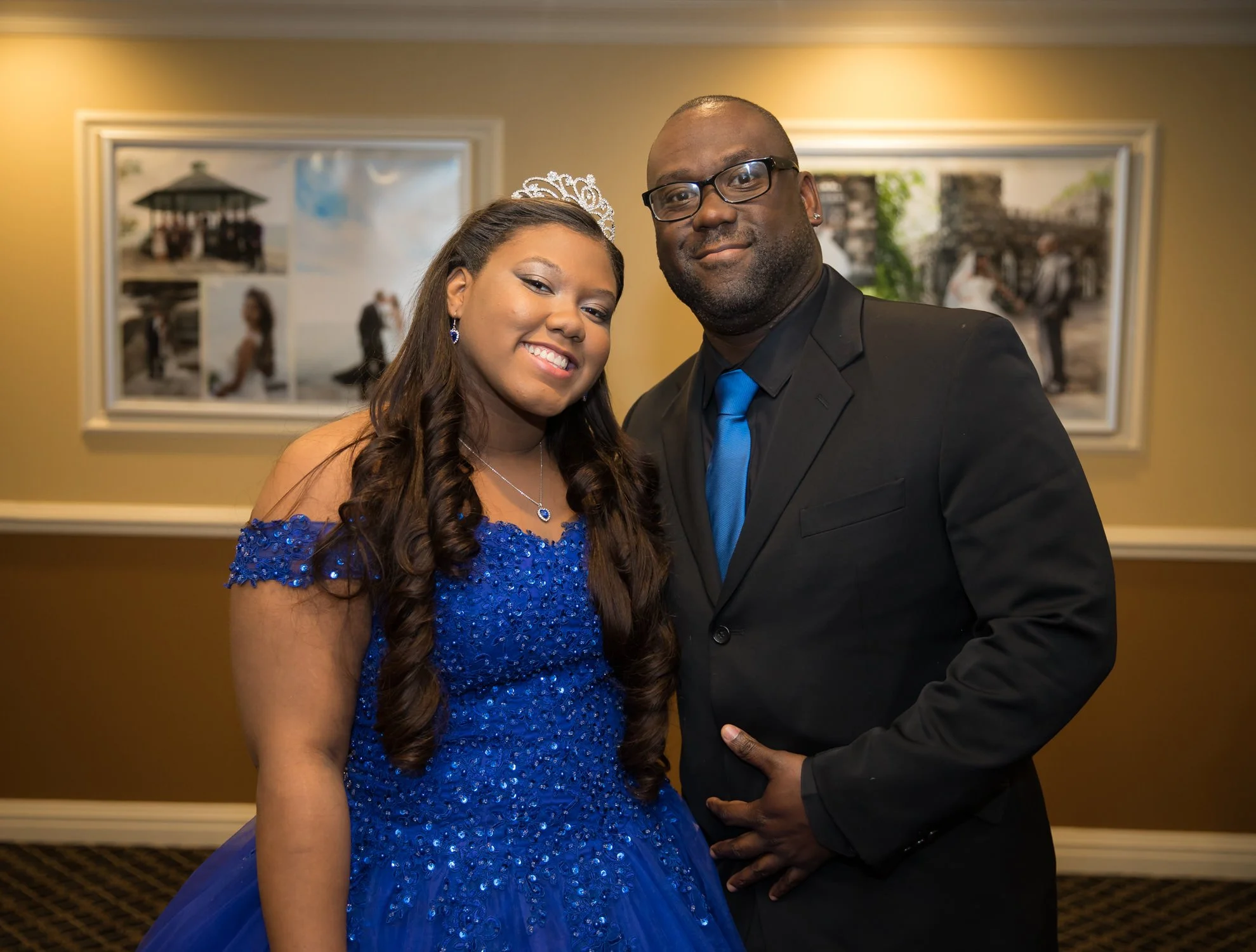 A young woman wearing a blue dress and a tiara, standing next to a man in a black suit, in an indoor setting with framed photos on the wall behind them.